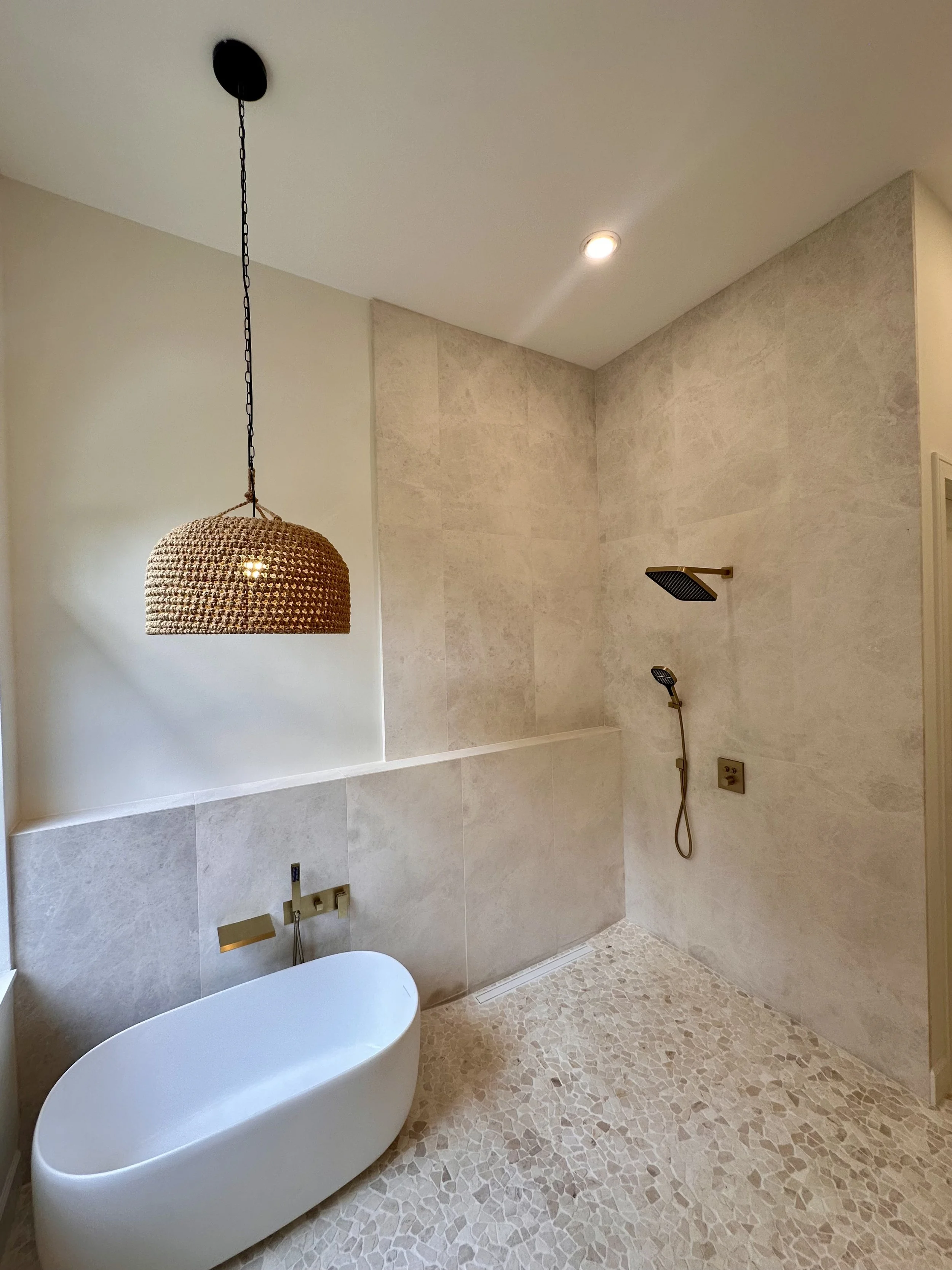 A modern bathroom with a freestanding oval bathtub, a black rain showerhead, and a handheld shower attachment inside a tiled shower area with a pebble floor. There is a woven pendant light hanging from the ceiling and recessed lighting.