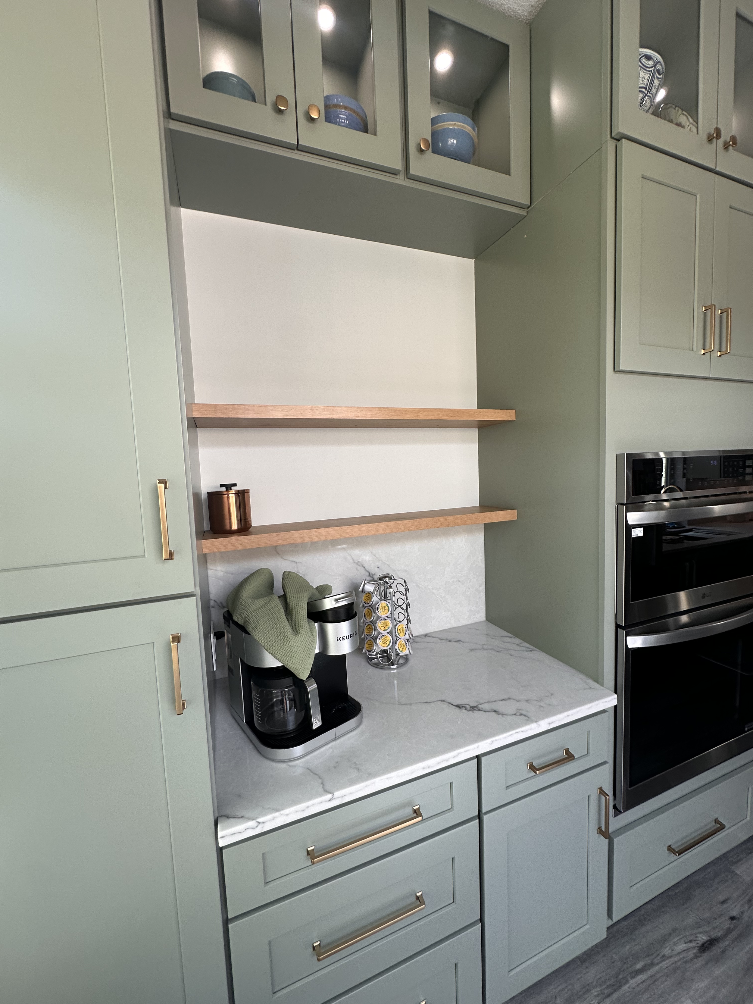 A modern kitchen with light green cabinets, white marble countertop, wooden open shelves, and a coffee machine with a green oven mitt on top. The kitchen features a built-in oven, microwave, and decorative bowls in glass-front cabinets.