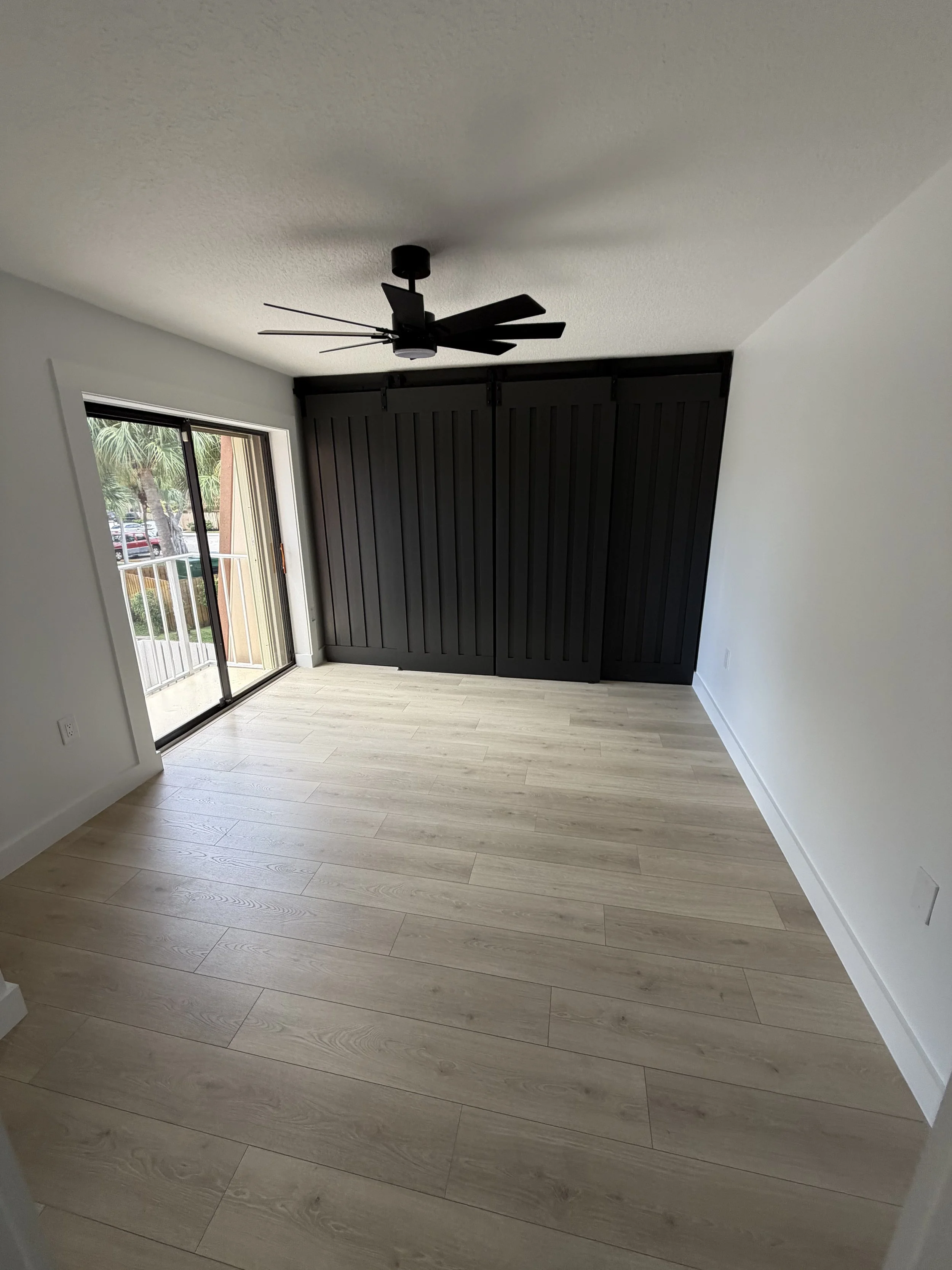 A room with light wood laminate flooring, white walls, a black ceiling fan, and a sliding glass door leading to a balcony with a view of palm trees and parked cars outside.