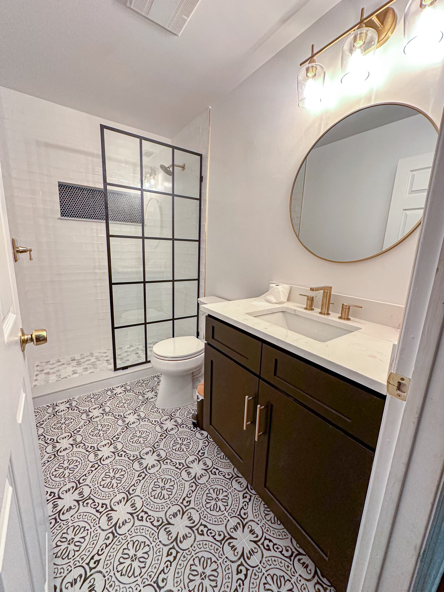 A modern bathroom featuring a walk-in shower with black metal framing, white subway tile, and hexagon black and white mosaic floor. The vanity has a black cabinet, white marble countertop, and gold fixtures. A large oval mirror with a gold frame hang