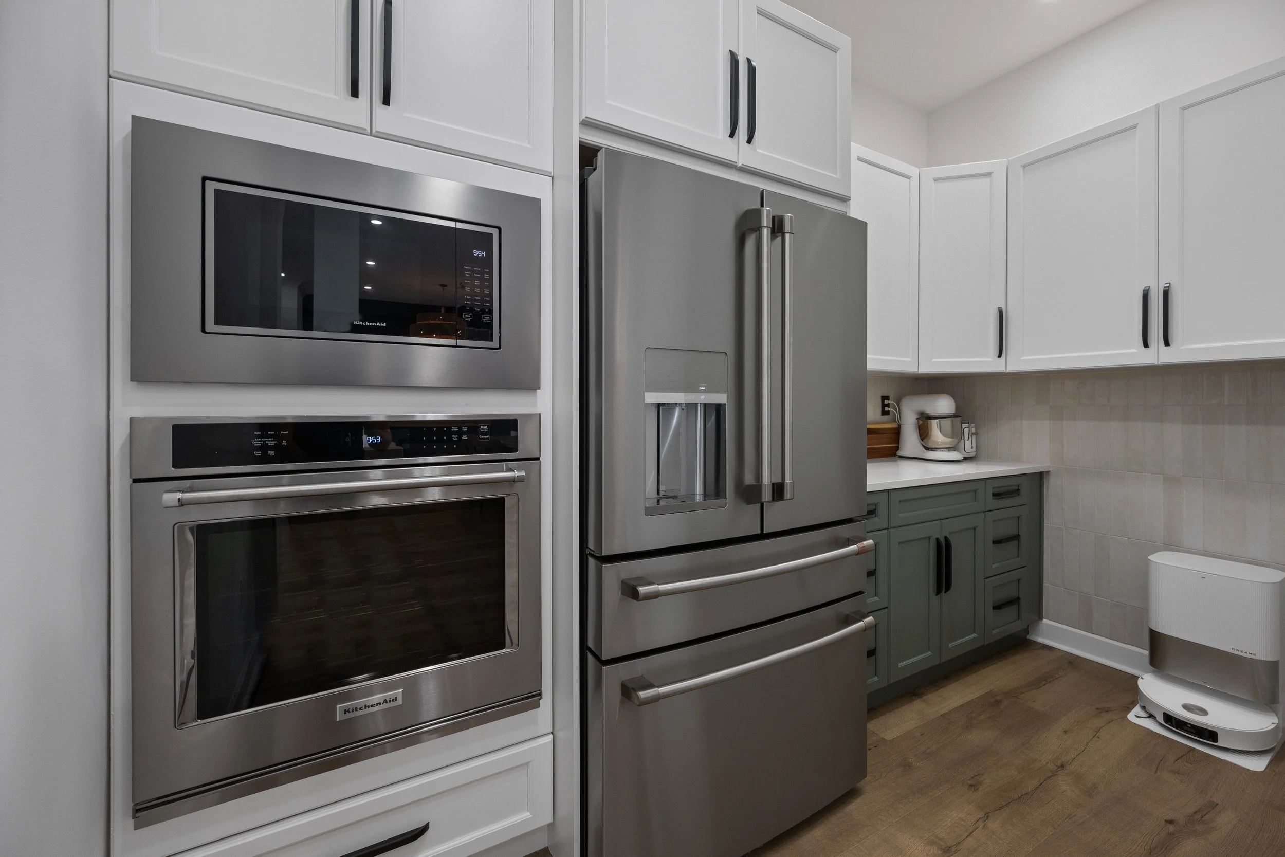 Modern kitchen with white cabinets, stainless steel microwave and oven, gray refrigerator, and a white countertop with a mixer. Floor is wood, and there are various appliances and storage areas.
