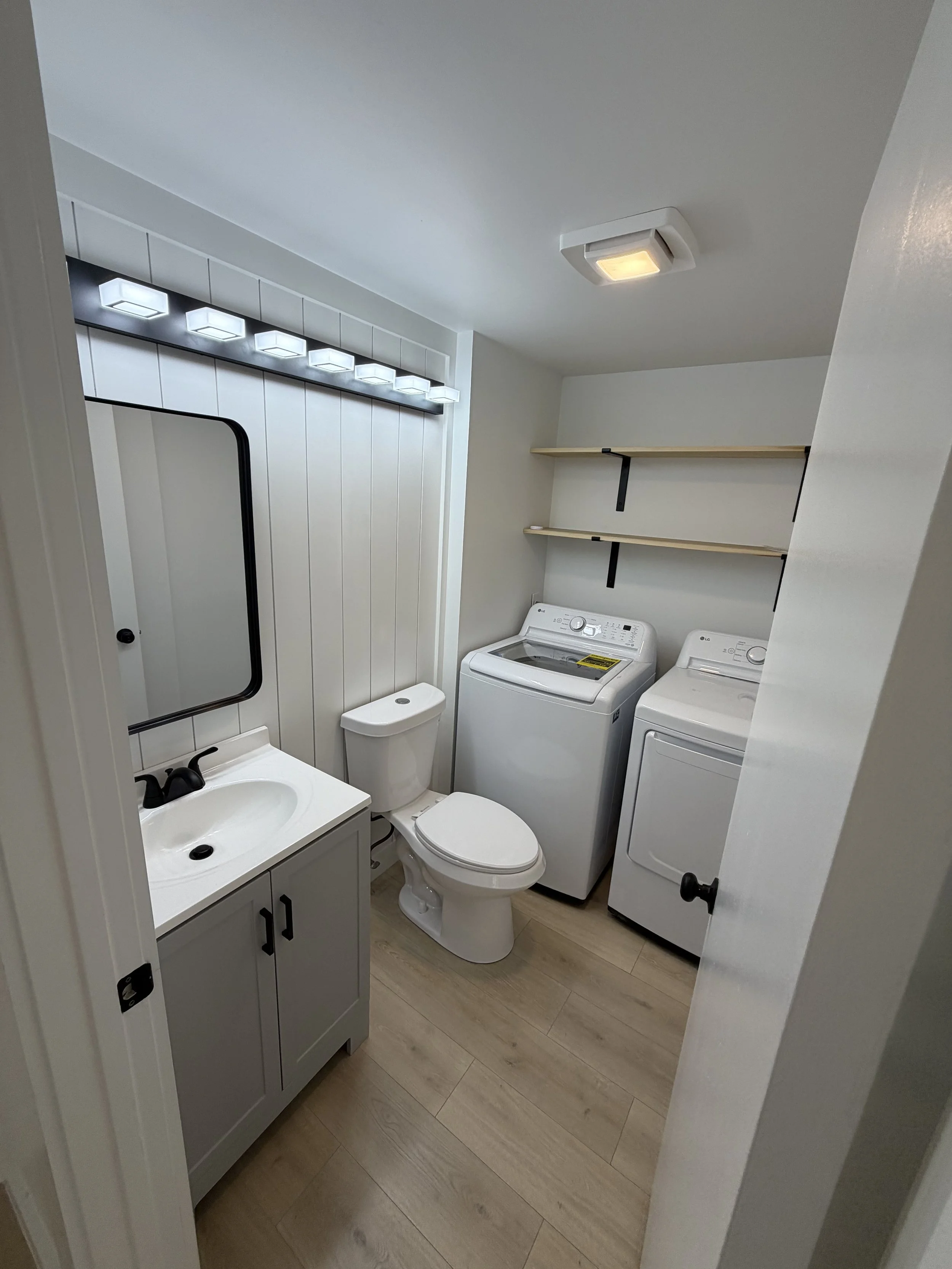 A small laundry room with a toilet, a sink vanity, a washing machine, a dryer, open shelving, and a ceiling light.