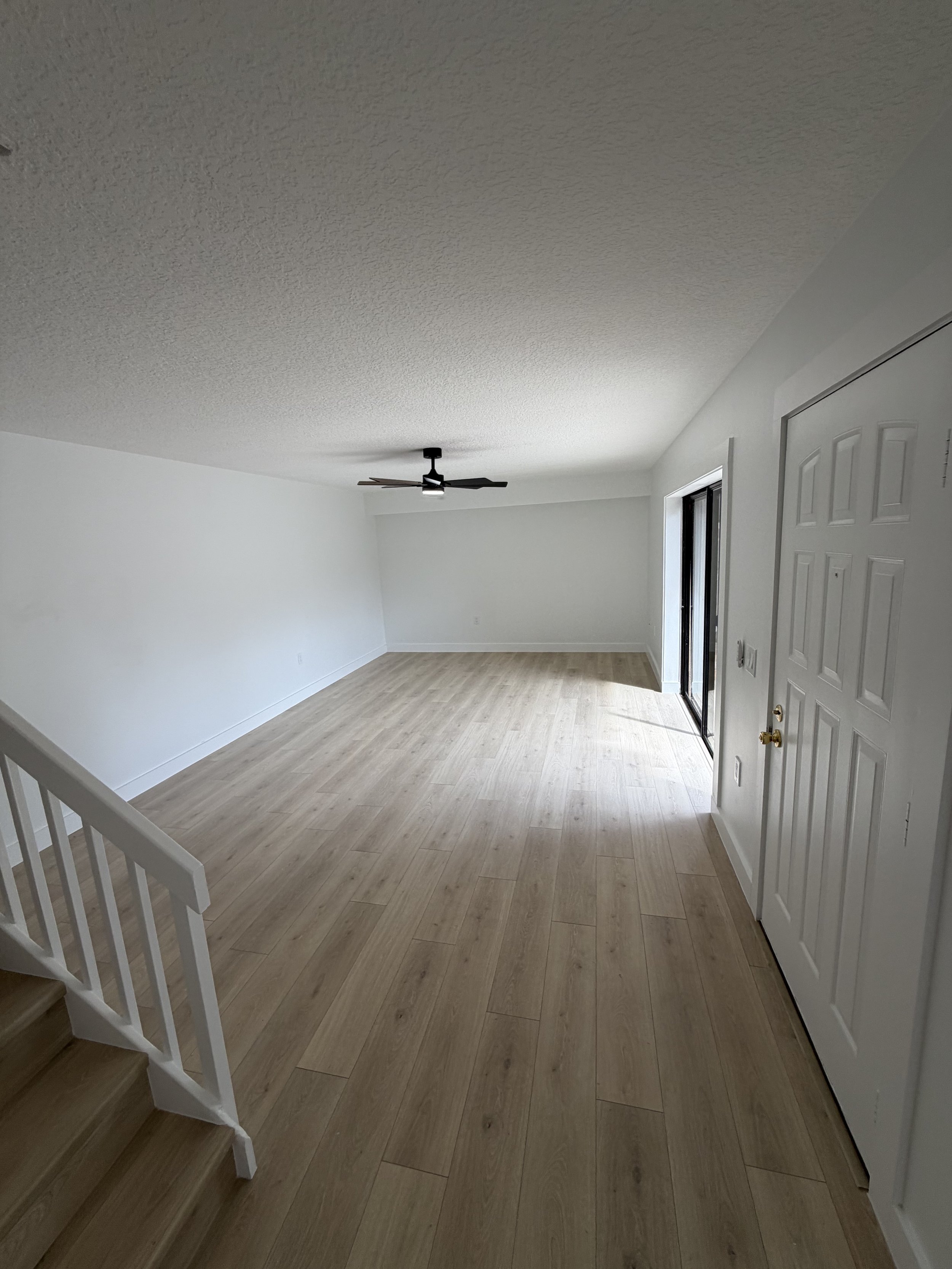 Empty living room with light-colored wood flooring, white walls, a black ceiling fan, a sliding glass door, and a white interior door.