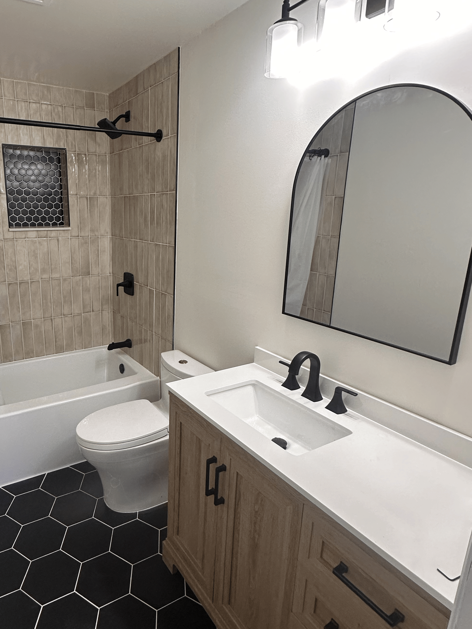 Modern bathroom with a white vanity, black fixtures, a large mirror, a toilet, and a shower area with beige tiles and a black showerhead.