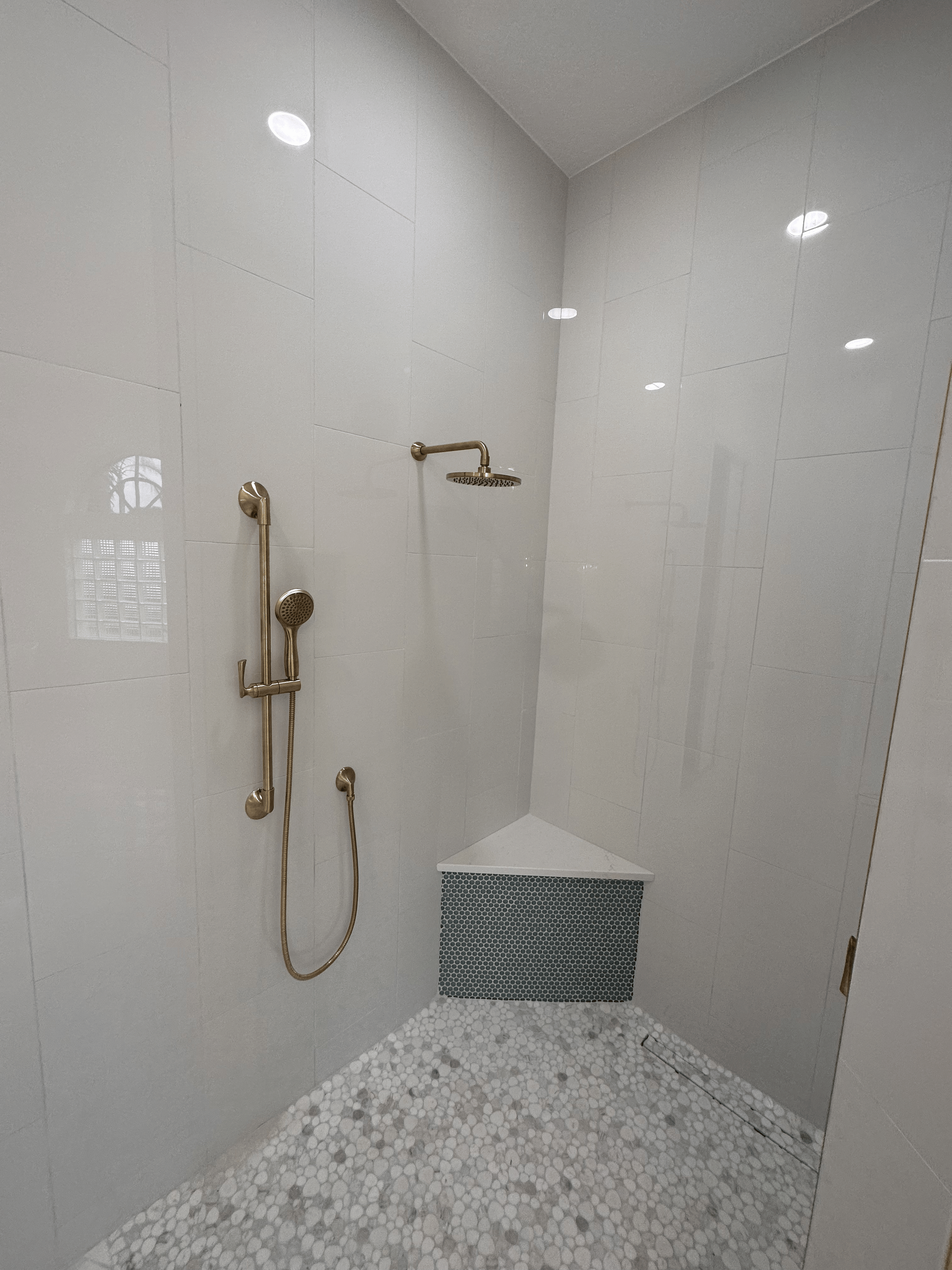 A modern walk-in shower with white tiled walls, a rain shower head, a handheld shower, and a pebble stone floor.