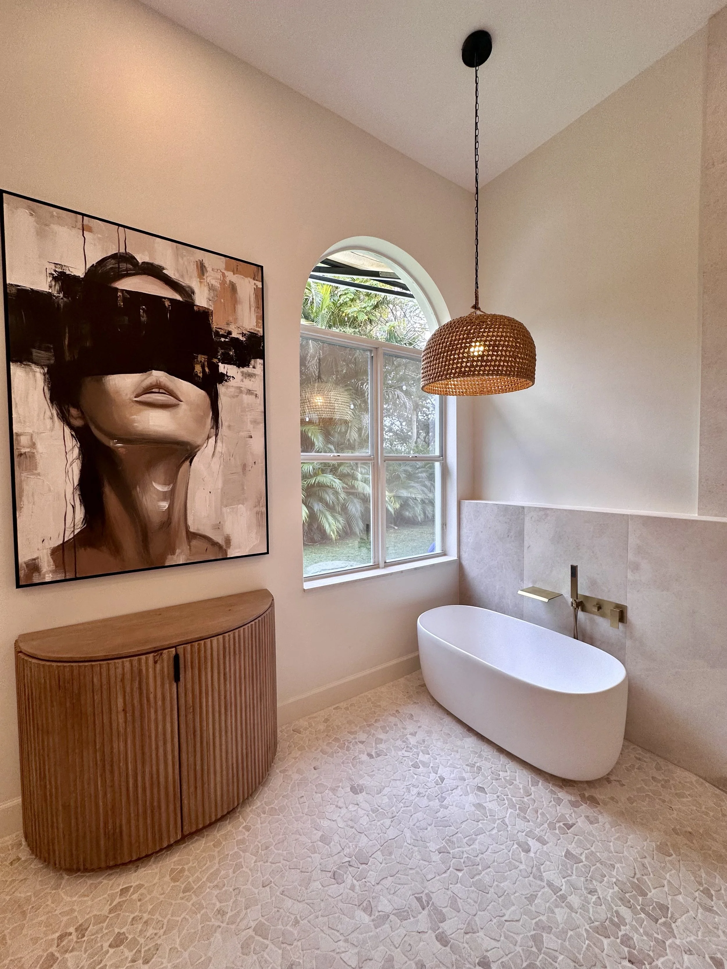 Modern bathroom with a white free-standing bathtub, a large window showing outdoor greenery, a hanging woven pendant light, a wooden cabinet with a curved design, and a portrait artwork on the wall.