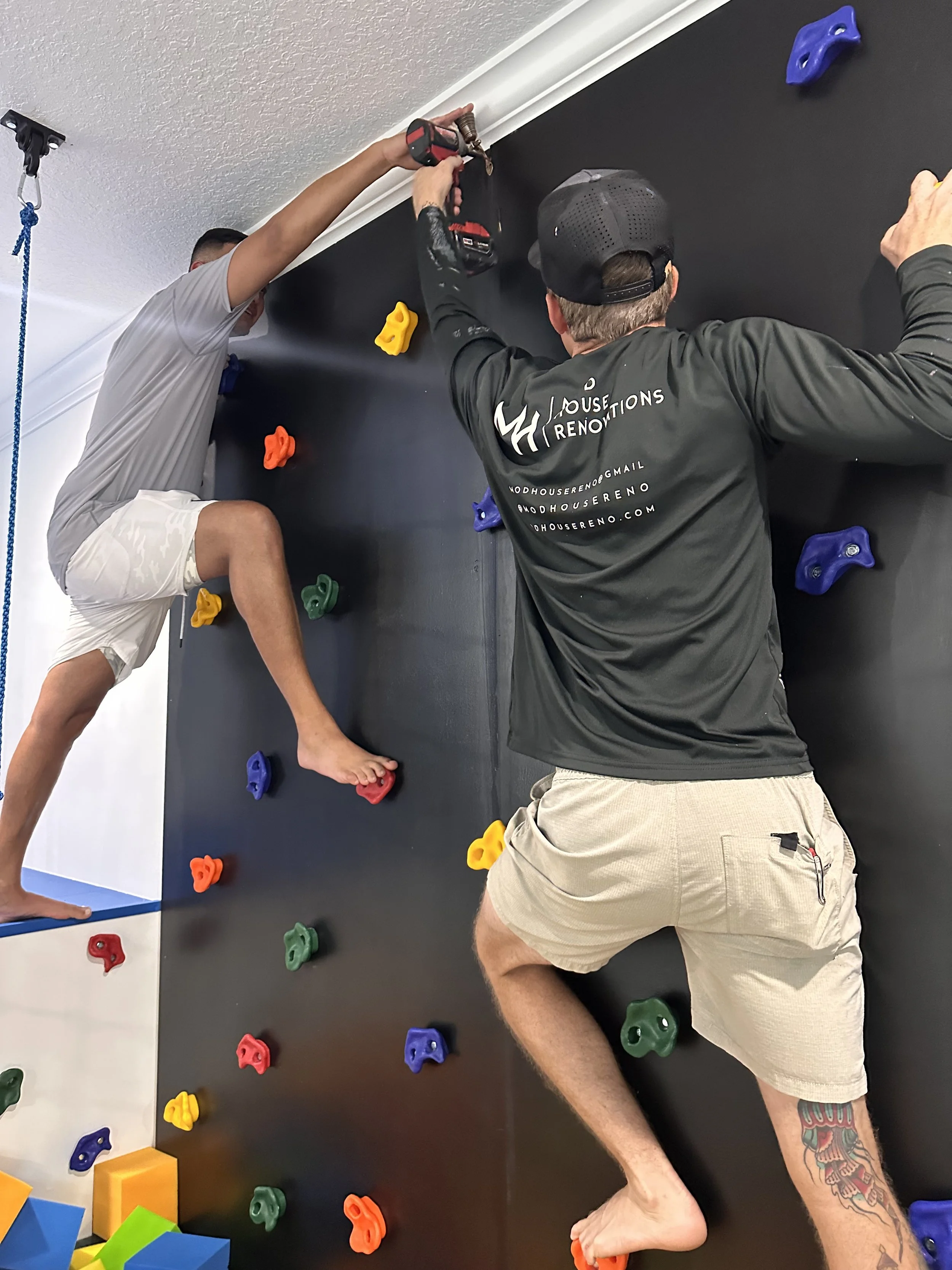 Two men are installing a black wall with colorful climbing holds. One man is using a drill, while the other is holding the wall steady. The climbing holds are in various colors, including yellow, red, green, purple, and blue. One man is wearing a gra