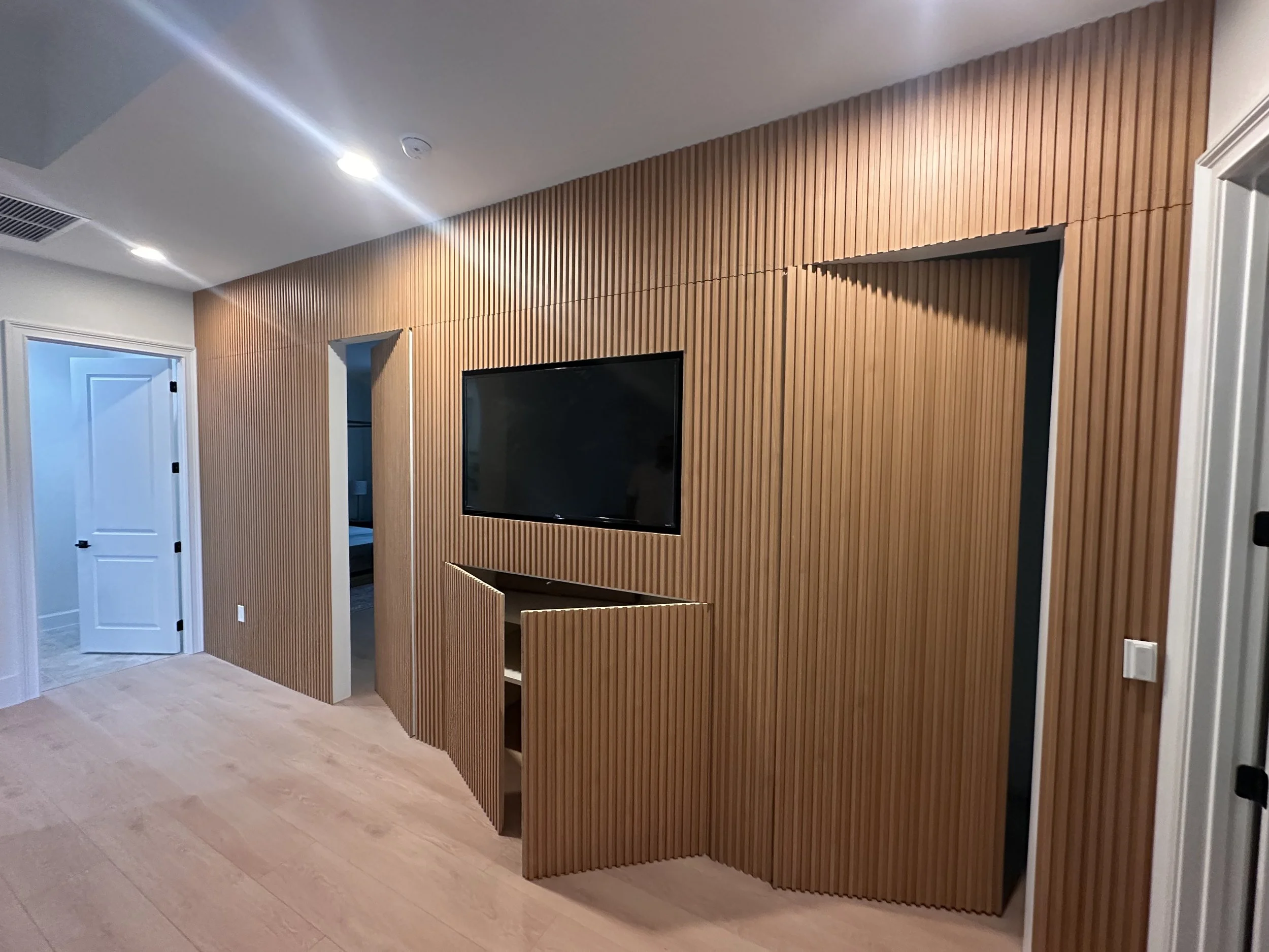 Living room with wooden slat wall, mounted flat-screen TV, white door on the left, and beige flooring.