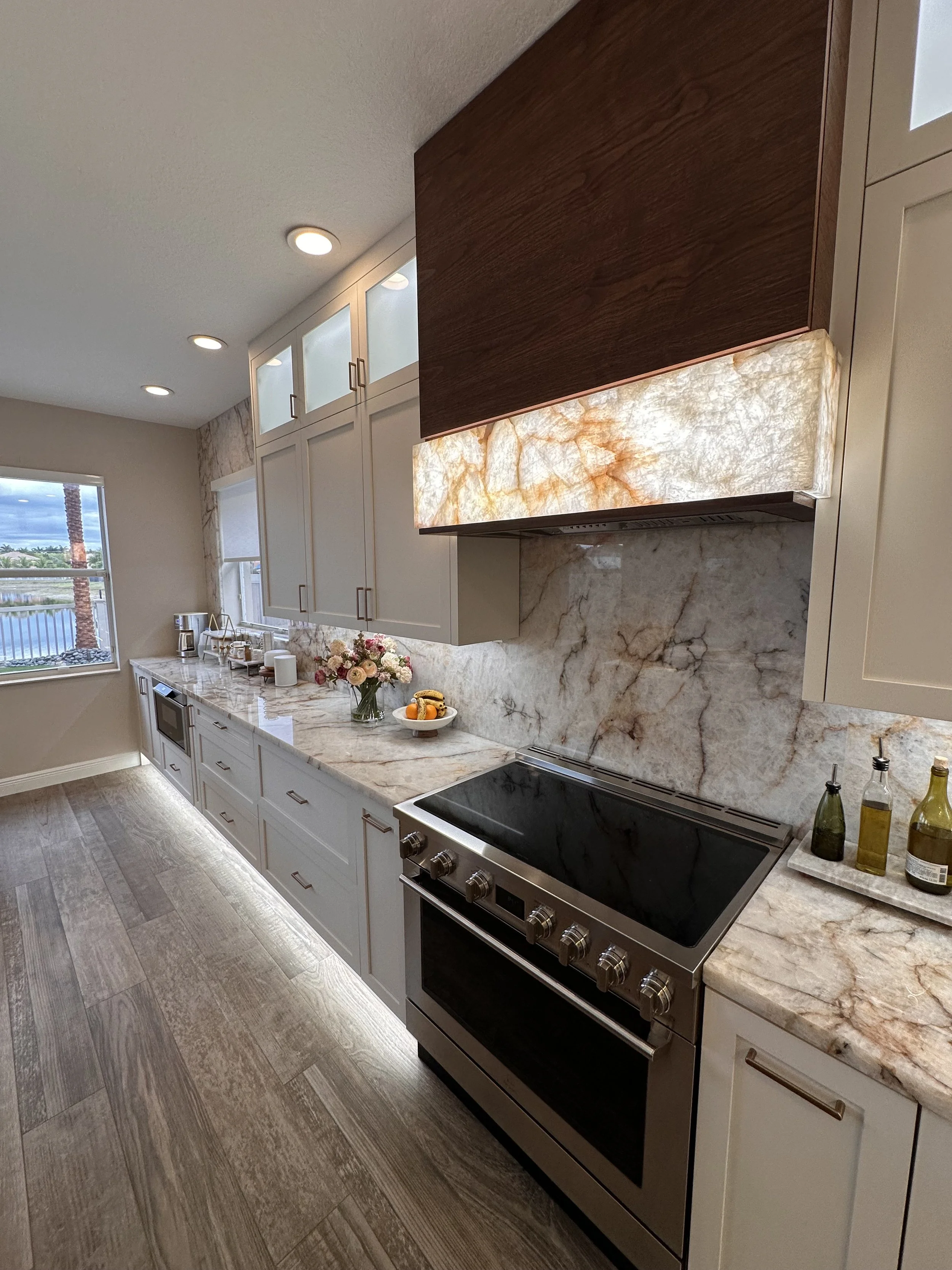 A modern kitchen with white cabinets, a marble backsplash, and a stainless steel stove. There are bottles of oil on the counter, a bouquet of flowers, and a plate of fruit. The kitchen has large windows and overhead lighting.
