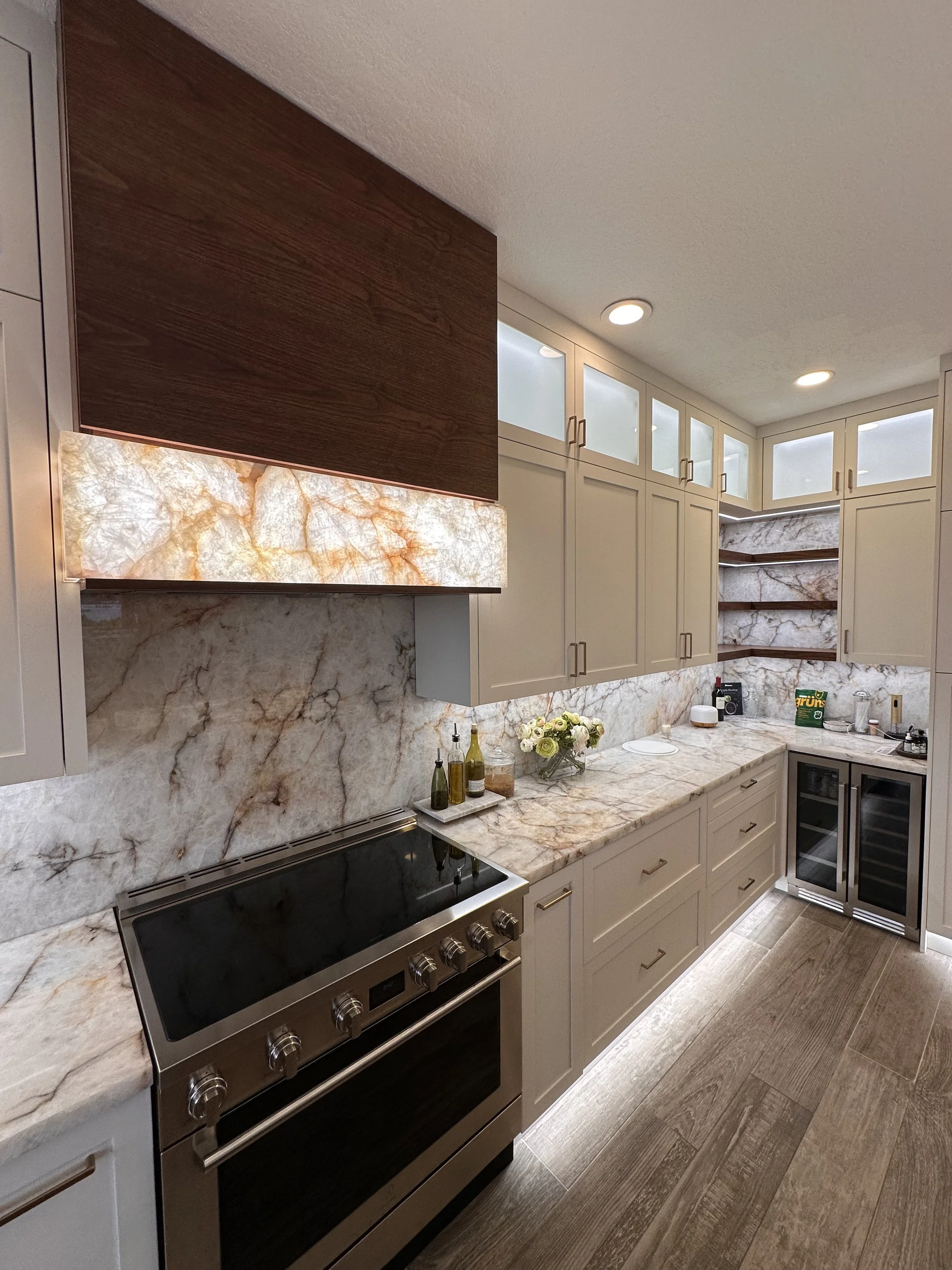 Modern kitchen with cream cabinets, a marble backsplash, and a marble countertop. Stainless steel stove and a wine cooler are visible, with decorative items like a flower arrangement and bottles on the counter.