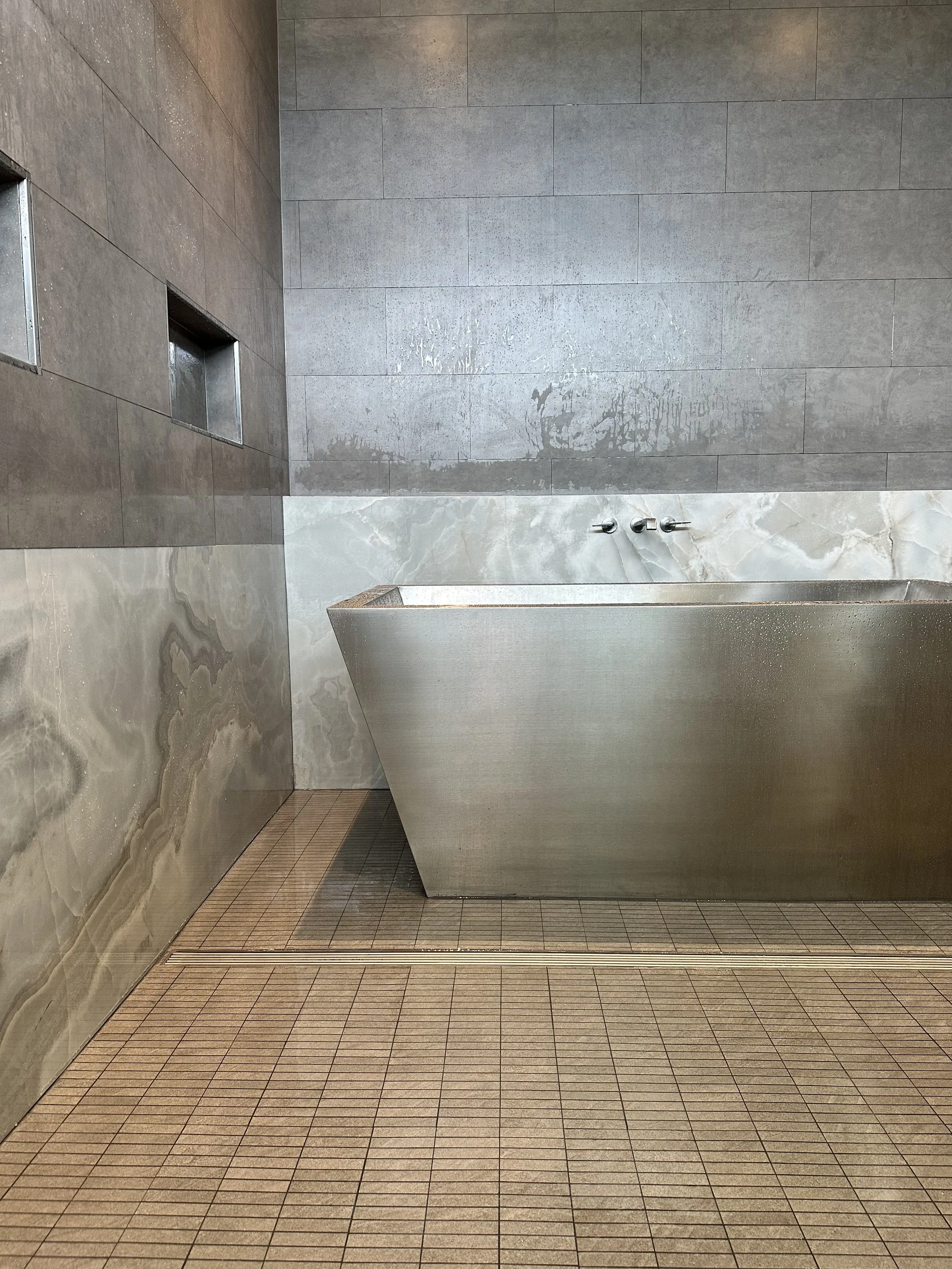 A modern public restroom shower stall with a tiled floor and walls, a marble partition, and water dripping from the ceiling pipe.