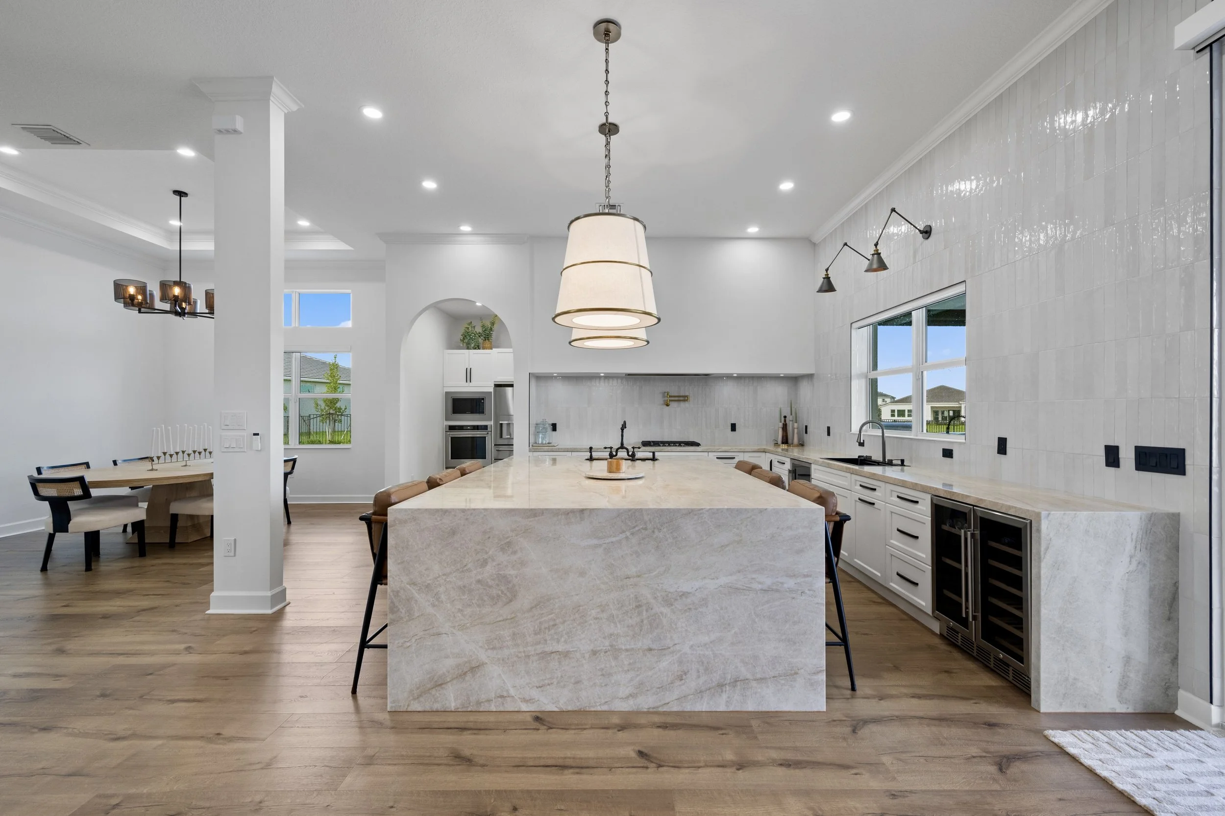 Open-concept kitchen with a large central marble island, white cabinetry, built-in microwave and fridge, and modern lighting fixtures. Adjacent dining area with a round table and black chairs is visible, with windows overlooking a backyard.
