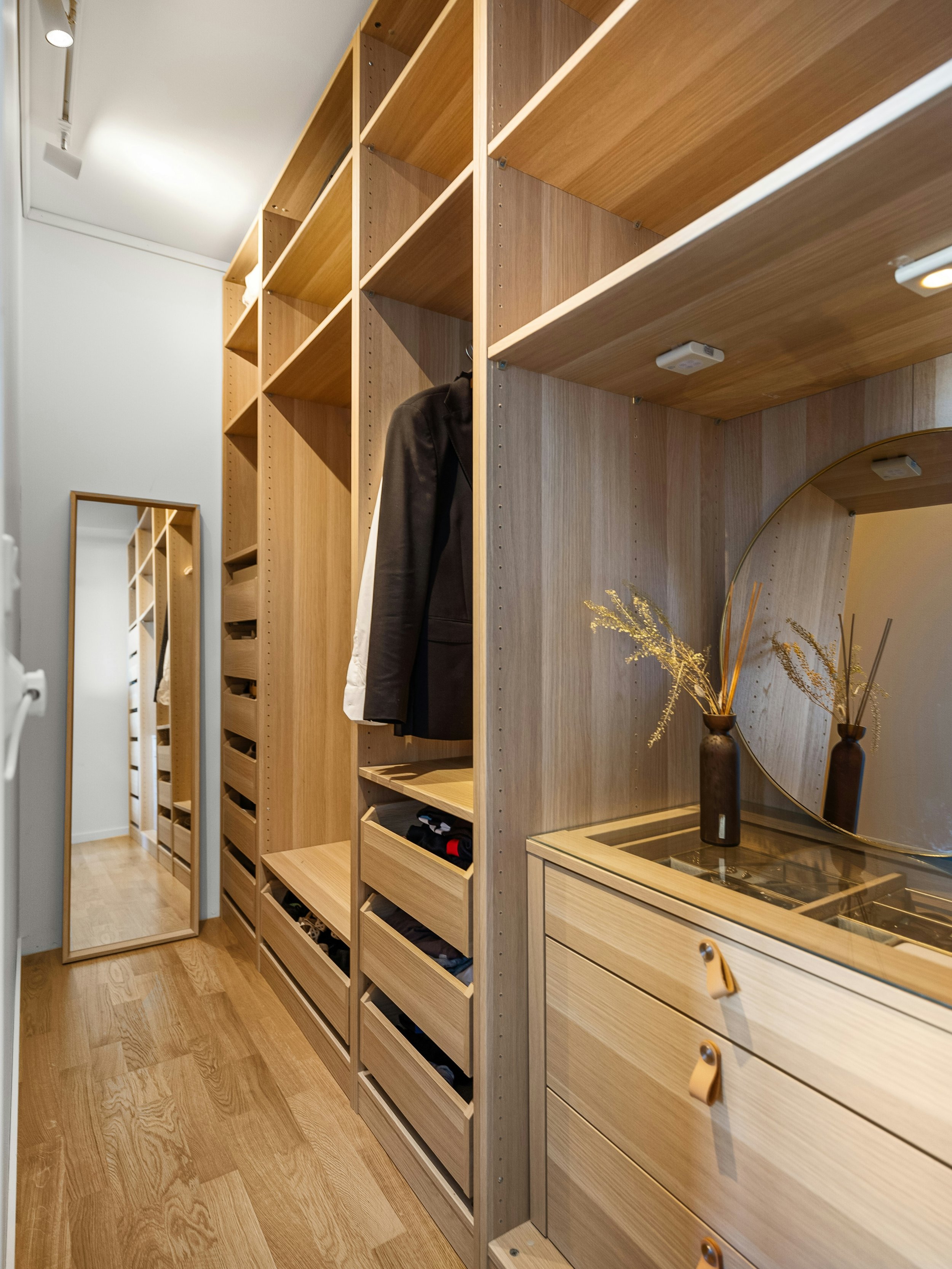 A modern wooden closet with open shelves, a mirror on the left, a black jacket hanging, and decorative vases with dried plants on a cabinet.