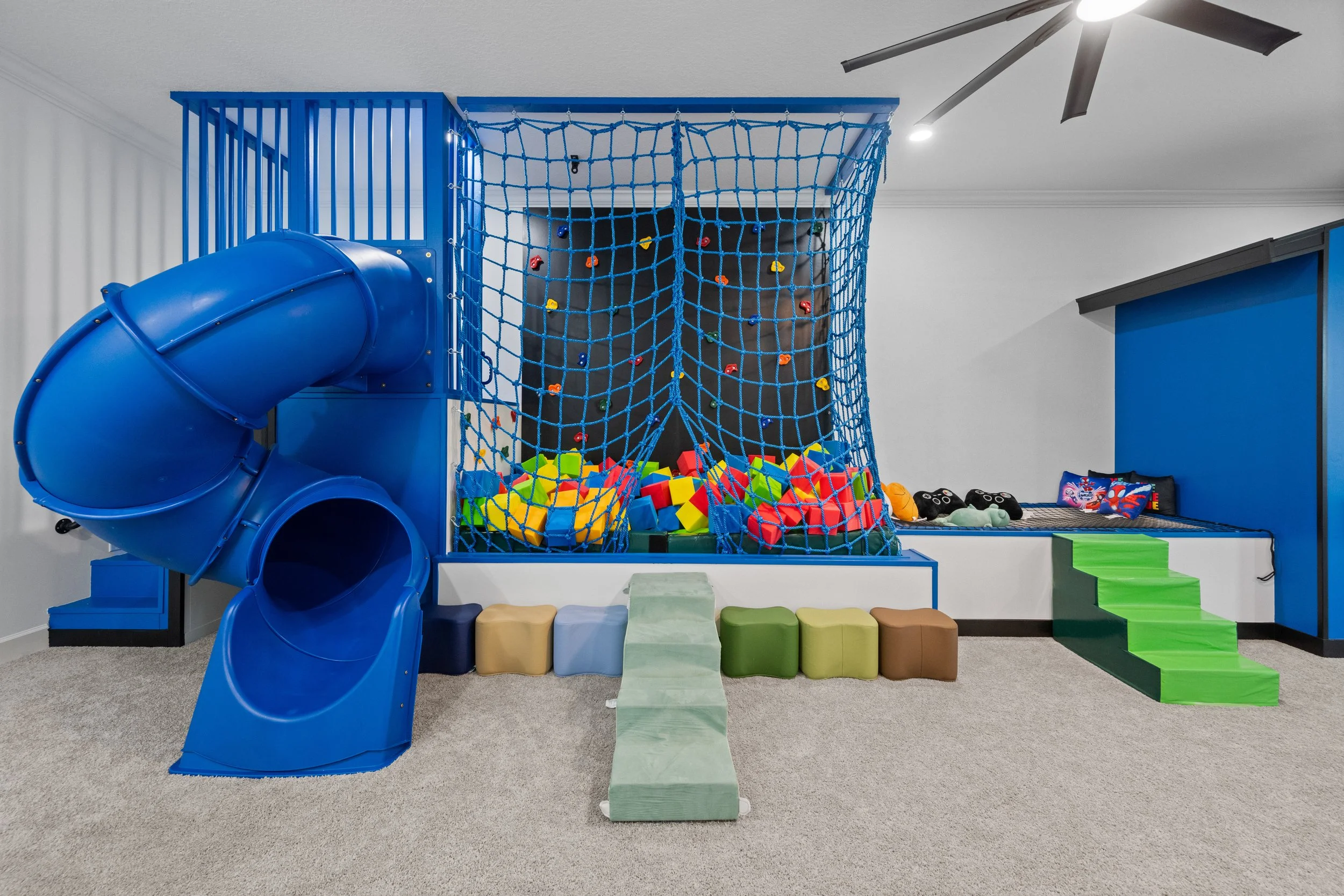 Indoor children's play area with a blue spiral slide, climbing net, foam blocks, and soft stepping stones on carpeted floor.
