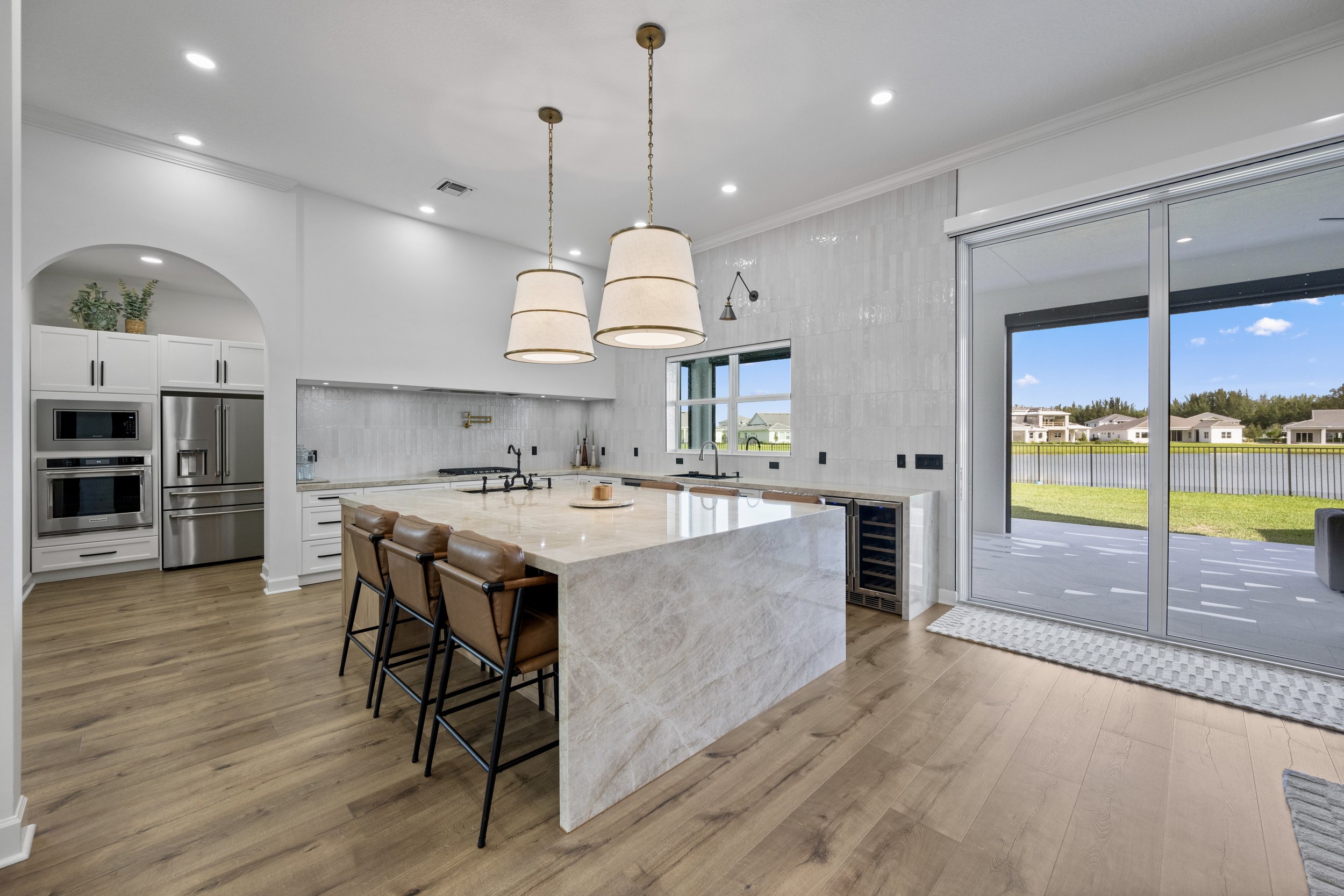Modern kitchen with a marble island, pendant lights, wood flooring, and large sliding glass door showing a backyard view.