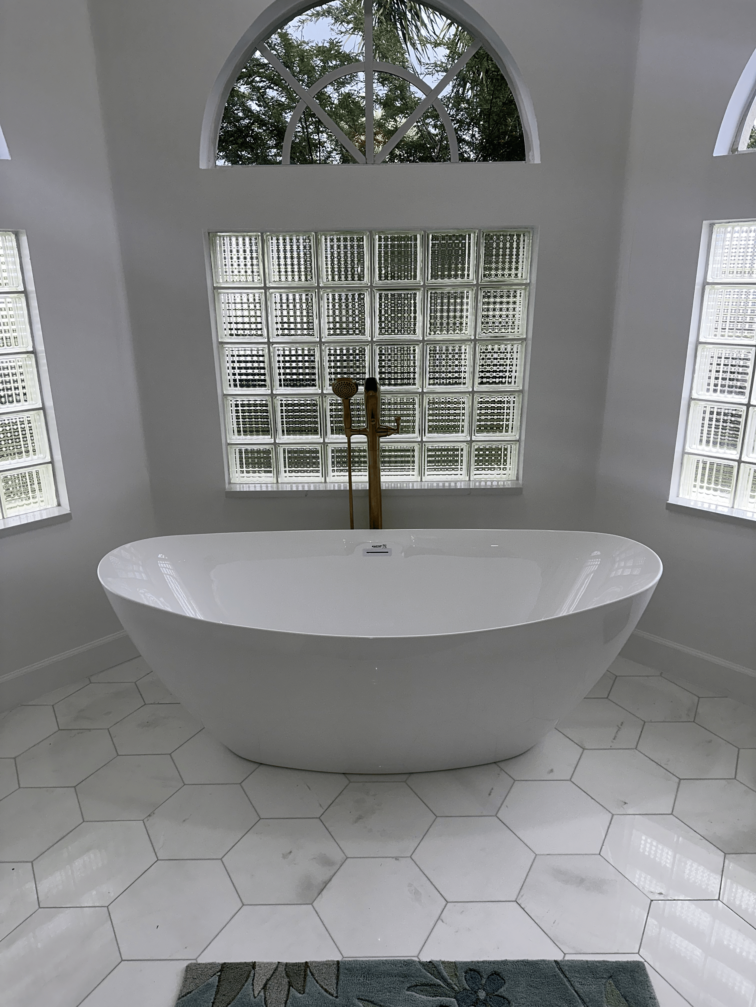 A white freestanding bathtub in front of a wall with glass block windows and an arched window above, in a bathroom with white hexagonal tile flooring and a grey rug.