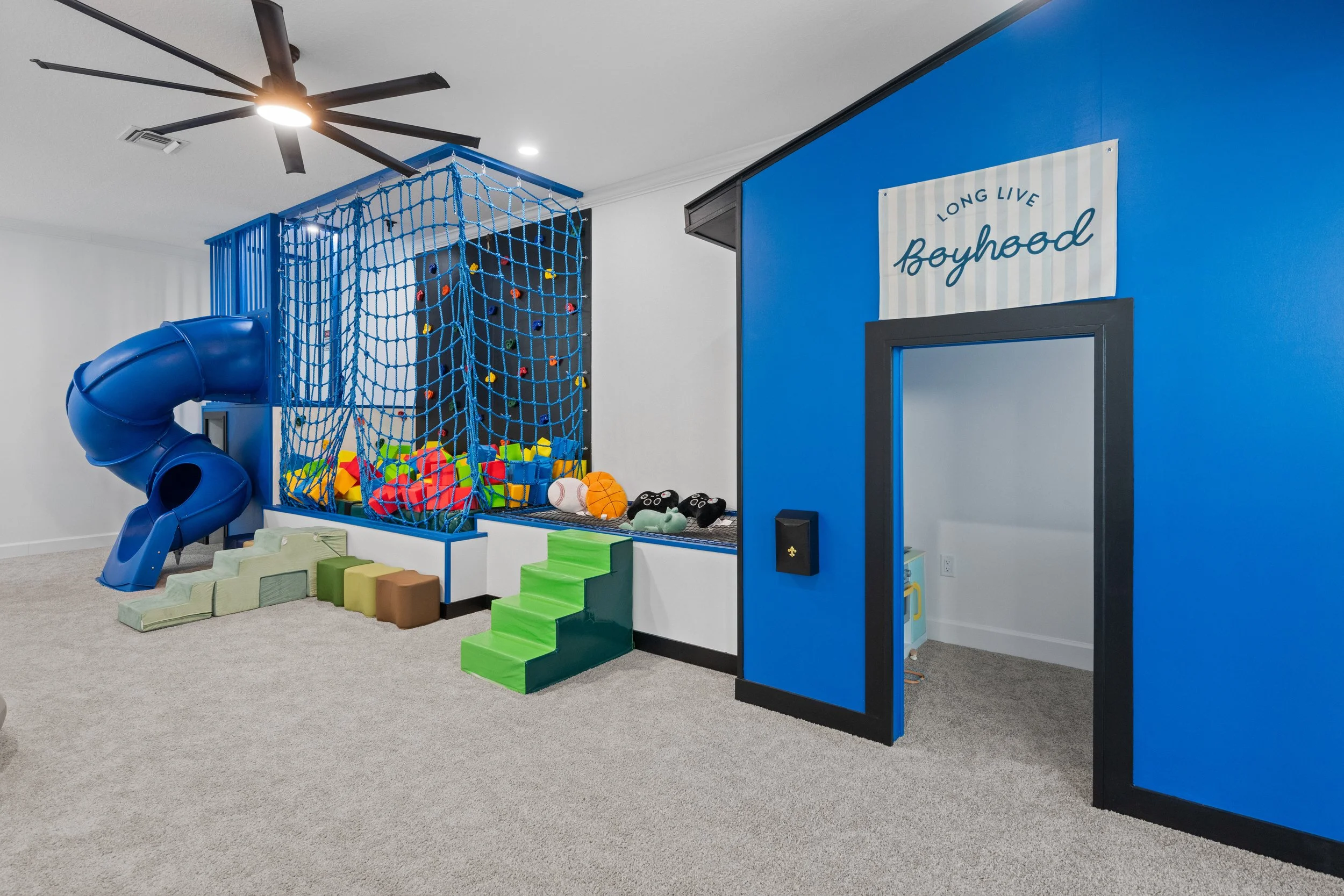 Indoor children's play area with a blue slide, climbing net, foam blocks, and a sign that reads 'Long Live Boyhood' on a blue wall.