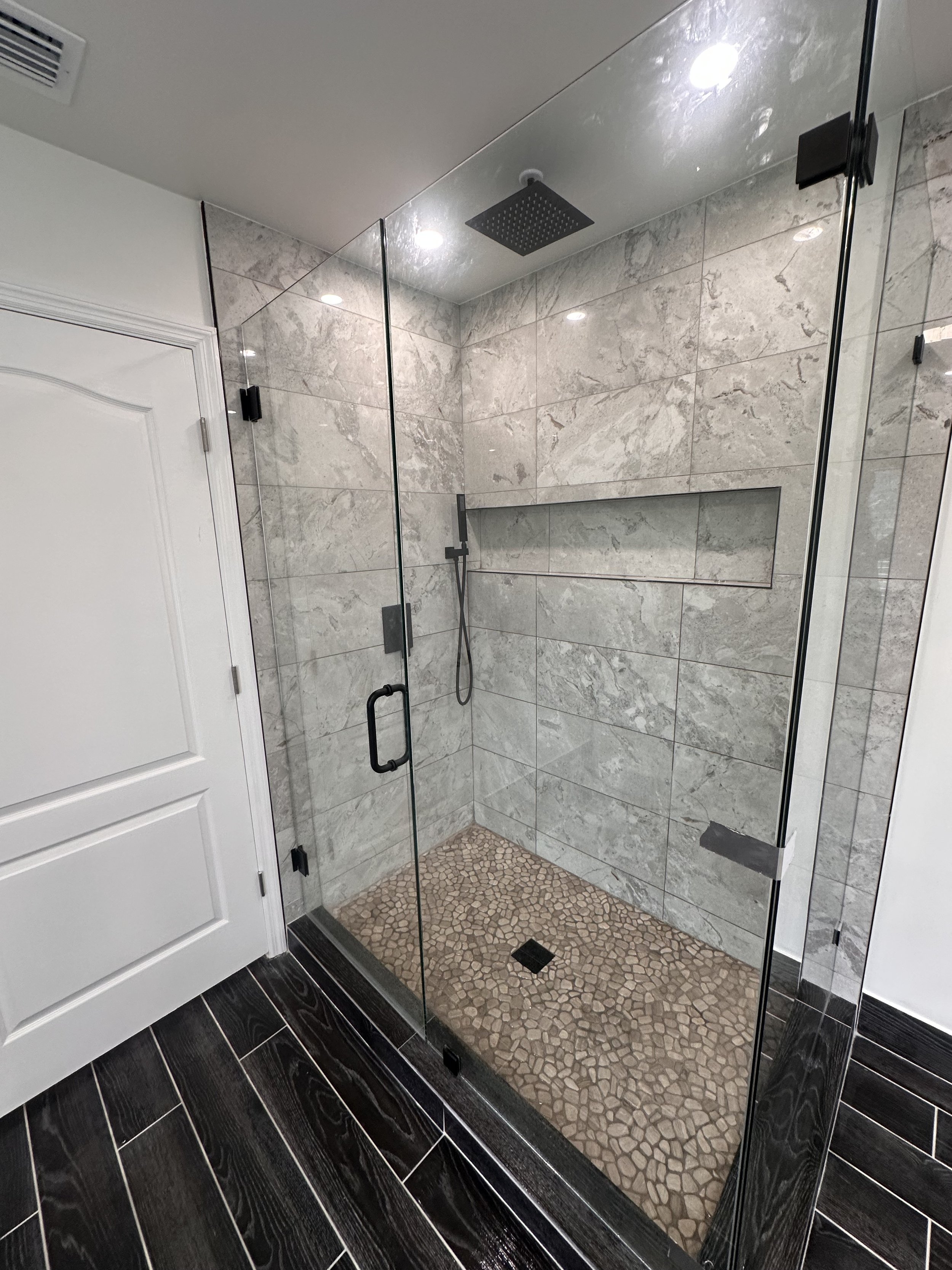 A modern walk-in shower with glass doors, beige and gray marble wall tiles, a black square rain shower head mounted on the ceiling, a handheld shower wand, a built-in wall niche, and a pebble tile floor, adjacent to a black tiled floor and a white do