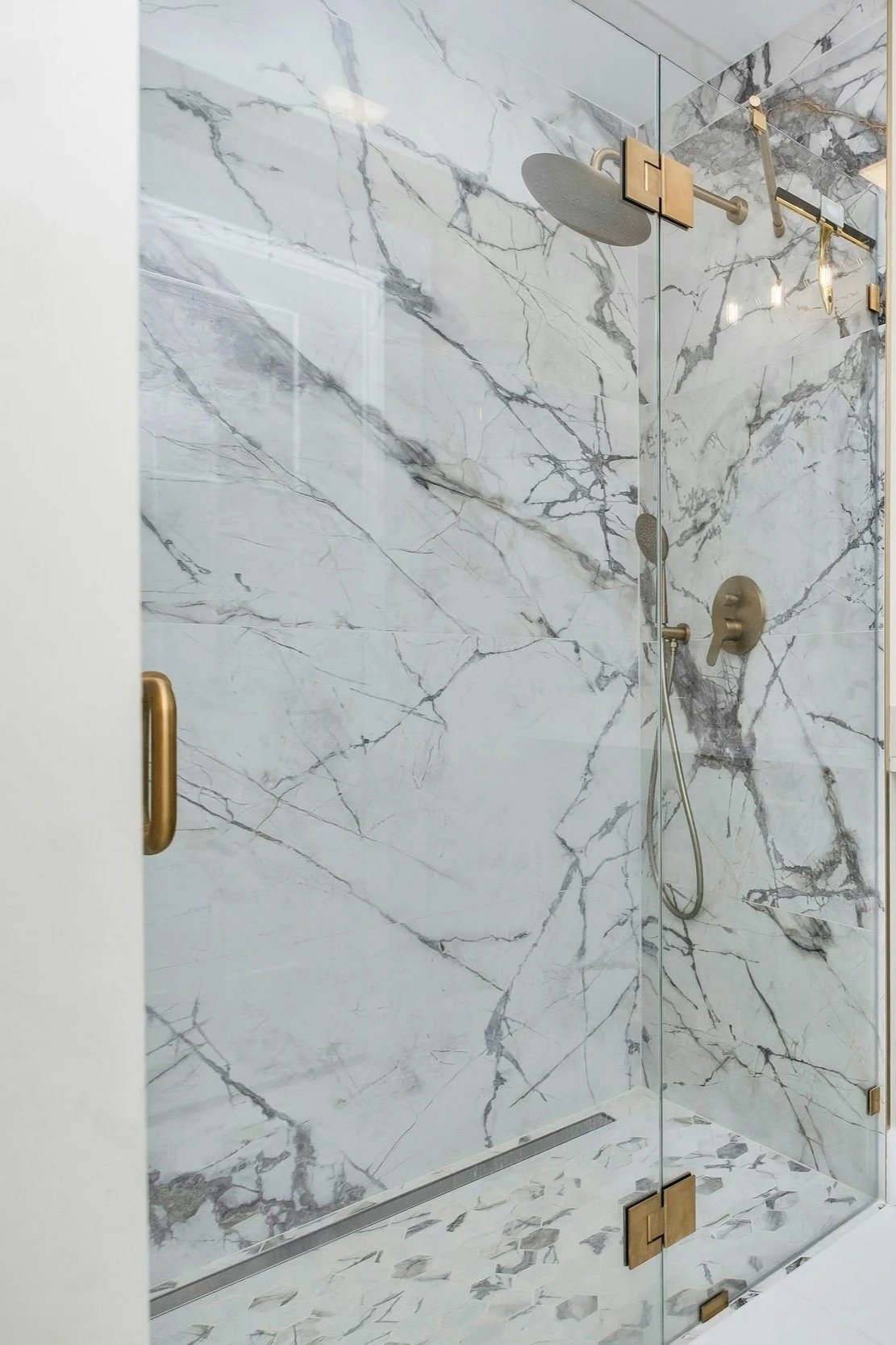 A marble shower with gold hardware, including a handheld showerhead, a large showerhead, and a glass door with gold hinges and handle.