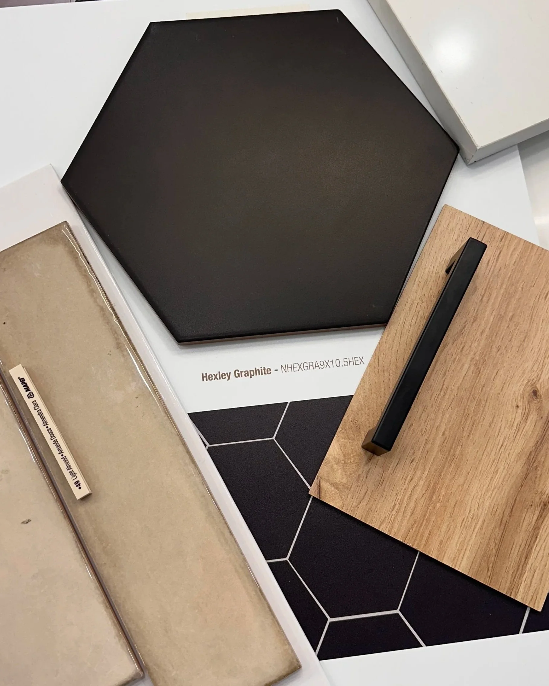 Samples of material and finishes including a black hexagonal tile sample, a beige material sample, and a wood grain sample with a black handle, displayed on a white surface.