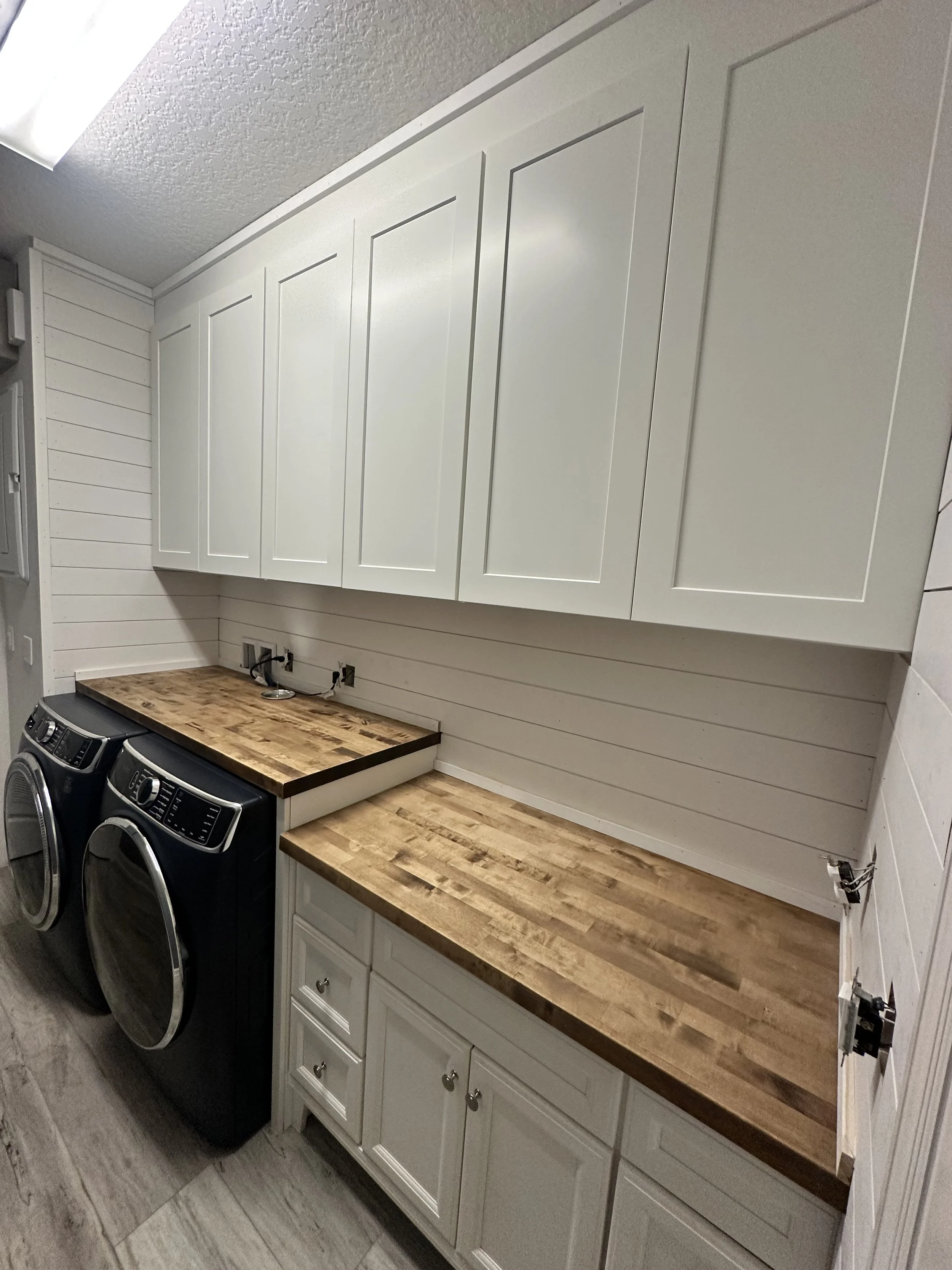 A laundry room with white cabinets, wooden countertops, and a V-shaped wall with hooks. Two front-loading washers are underneath the countertop. The wall has white horizontal wooden planks and electrical outlets.