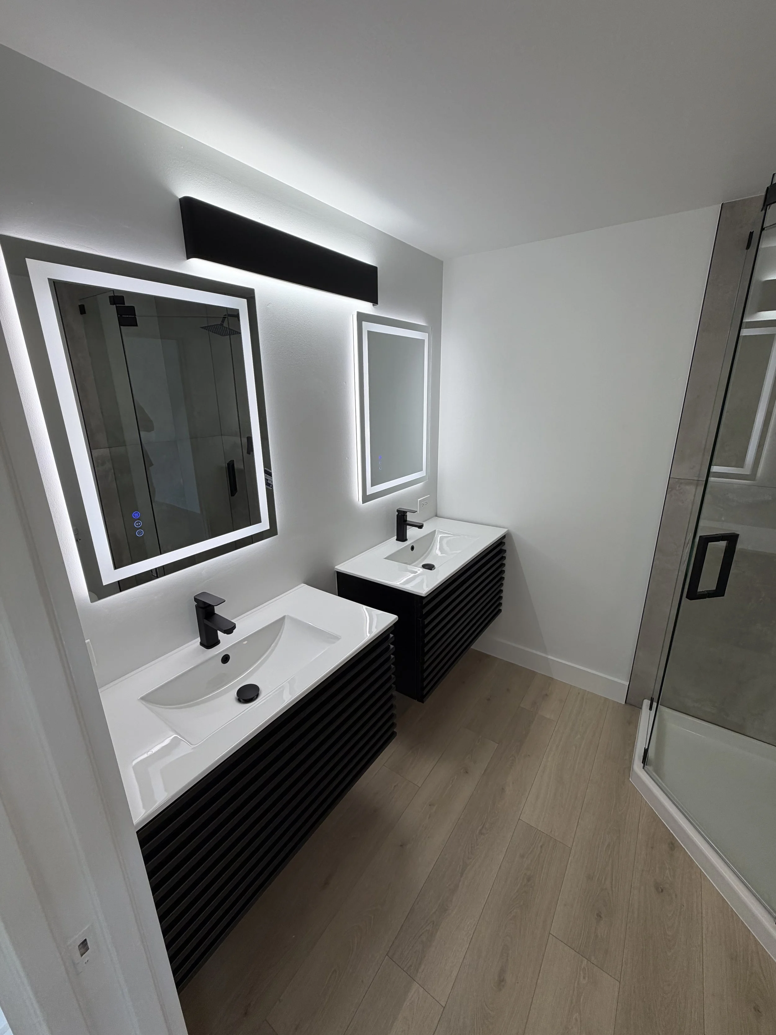 Modern bathroom with double sinks, illuminated mirrors, and a glass shower enclosure. The vanity has black horizontal slats, and the floor is wood.