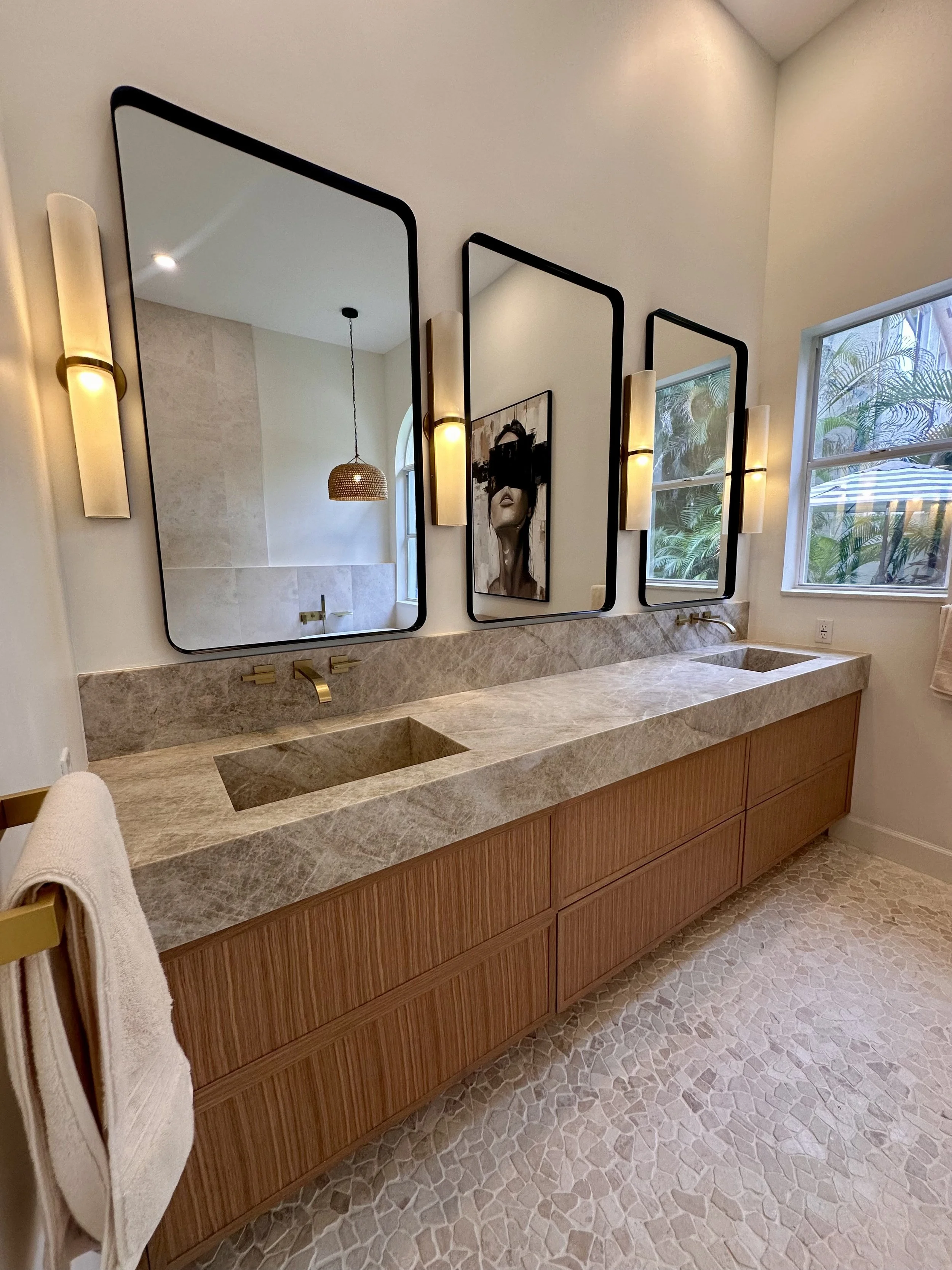 Bathroom vanity with a marble countertop, dual sinks, wood cabinet, large mirrors, wall lights, and a window with trees outside.