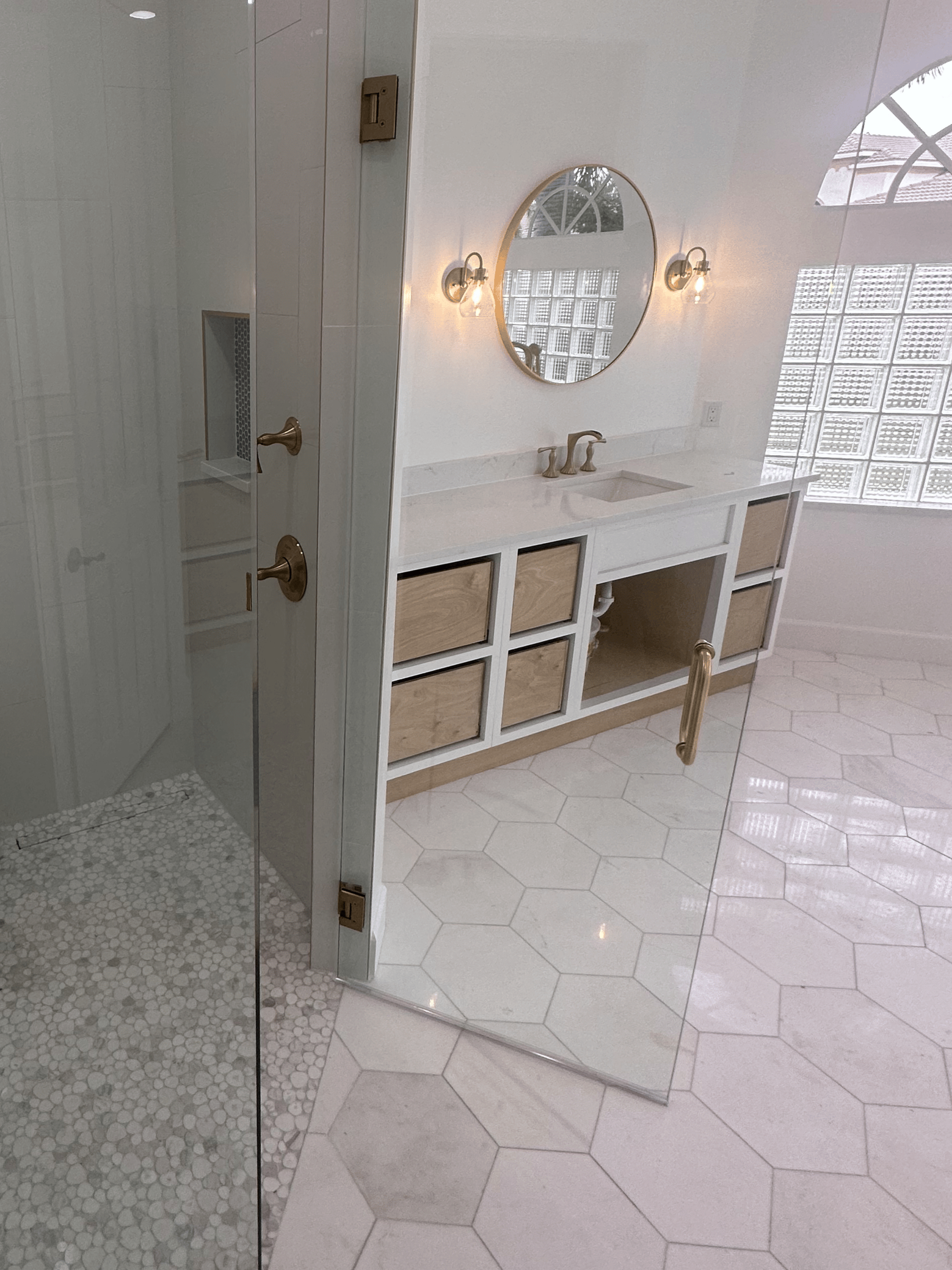 A bathroom with a glass shower door, a white vanity with a marble countertop, a round mirror, and wall-mounted light fixtures. There's a window with a curved top and a patterned glass block wall outside.