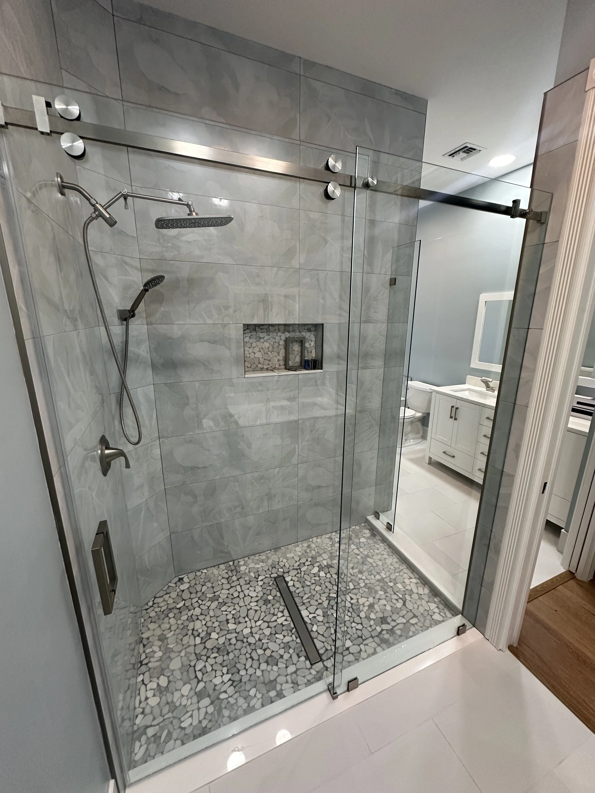 Modern bathroom with a glass shower enclosure, pebble stone shower floor, gray tile walls, and a built-in niche for toiletries. Visible mirror and toilet in the background.