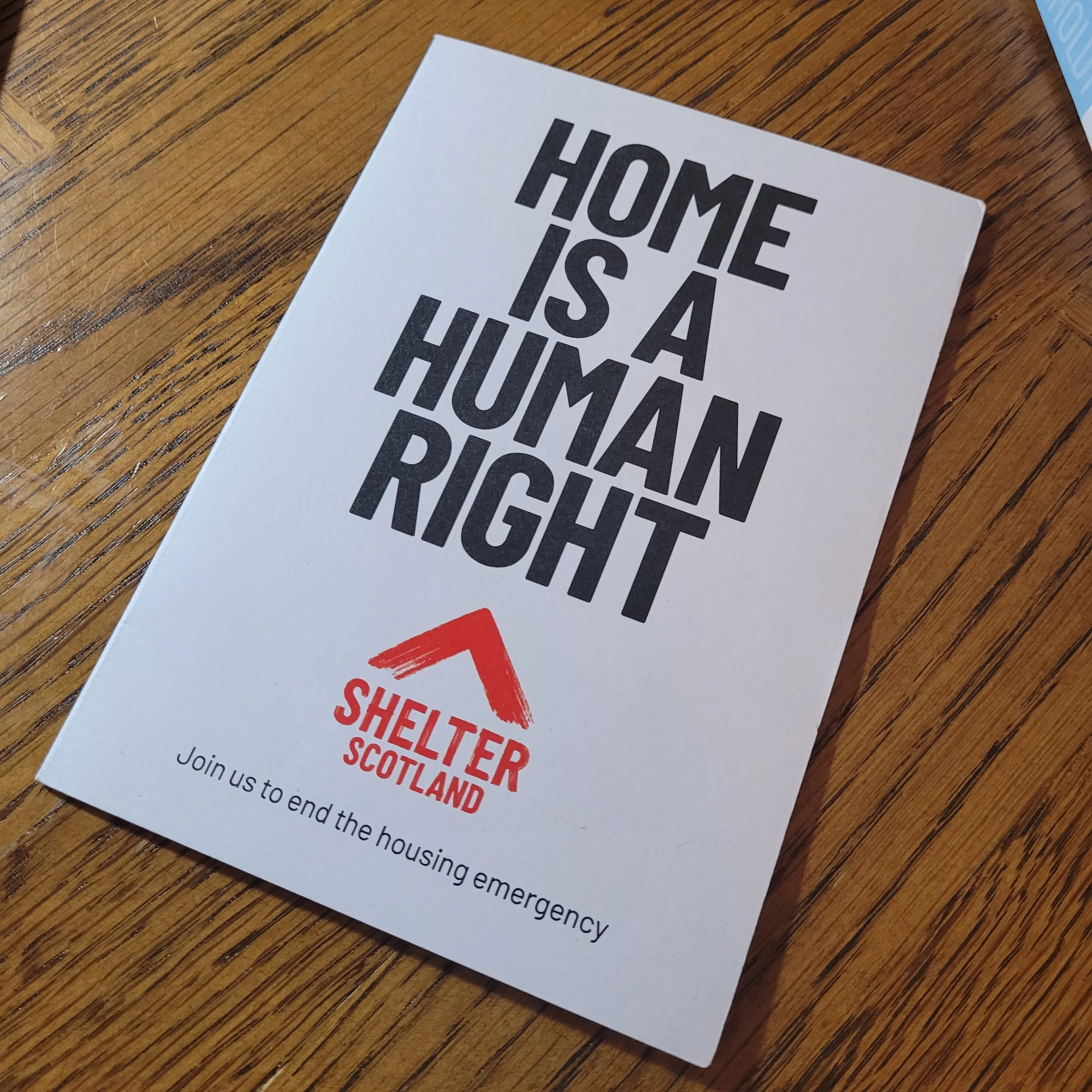 Leaflet on a wooden surface with bold text stating “Home is a Human Right,” the Shelter Scotland logo, and the message “Join us to end the housing emergency.”