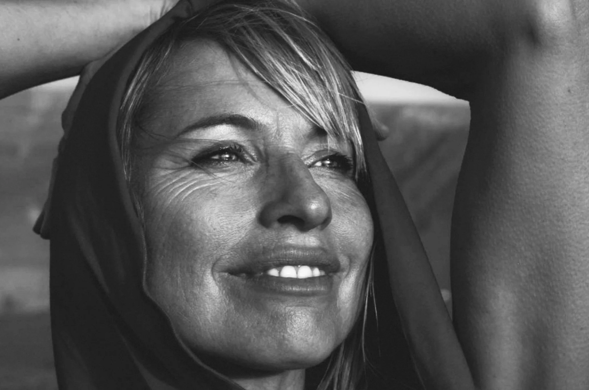 A black and white close-up of a woman smiling with her hand on her forehead, looking off into the distance.