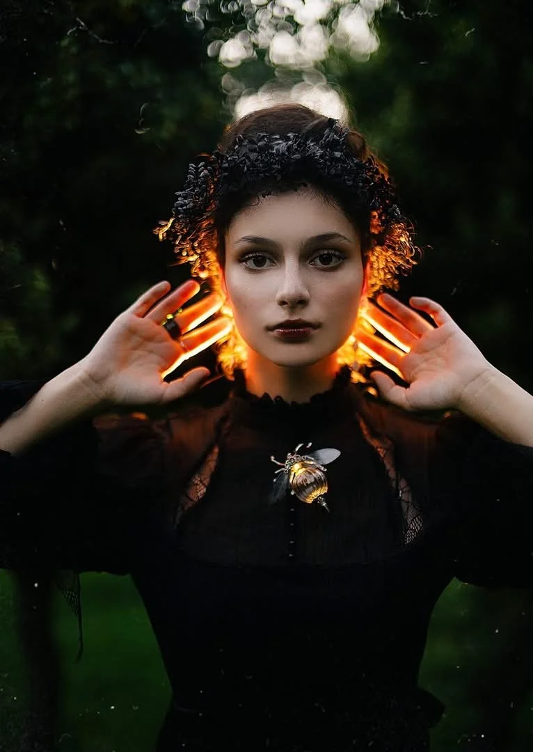 A young woman with dark makeup and curly hair wears a black headband and black dress, standing outdoors at dusk. She holds her hands near her head, with orange light illuminating her hair from behind, creating a halo effect. A large bee brooch is pinned to her dress.