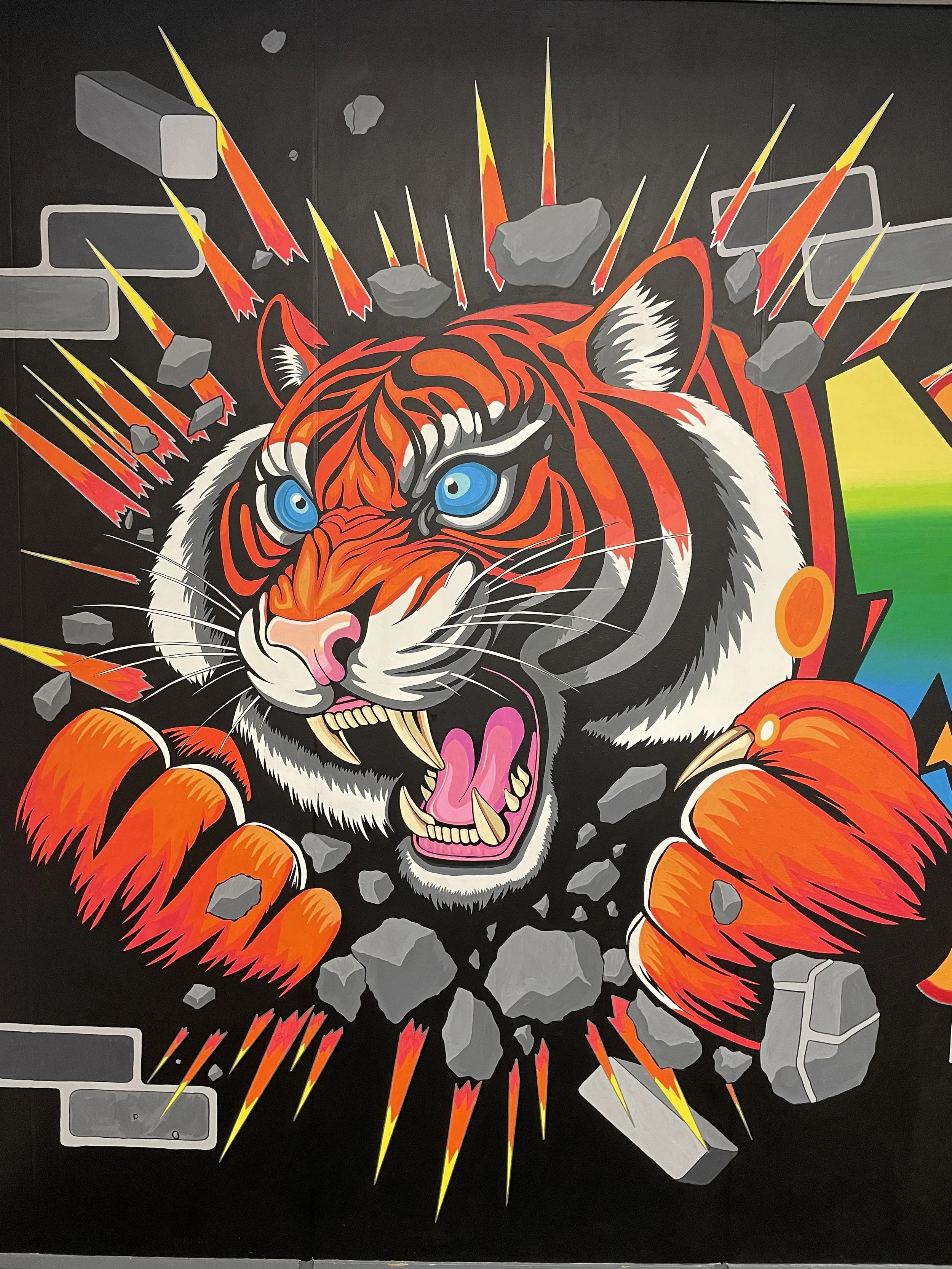 A vibrant graffiti mural of a roaring tiger breaking through a brick wall, with rocks and lightning effects around it.