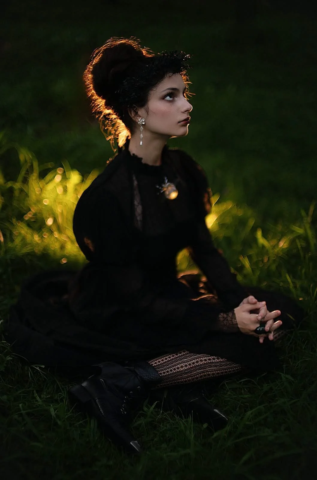 A woman dressed in gothic fashion, sitting on the grass during sunset, with dark makeup, lace clothing, and jewelry, illuminated by golden light.