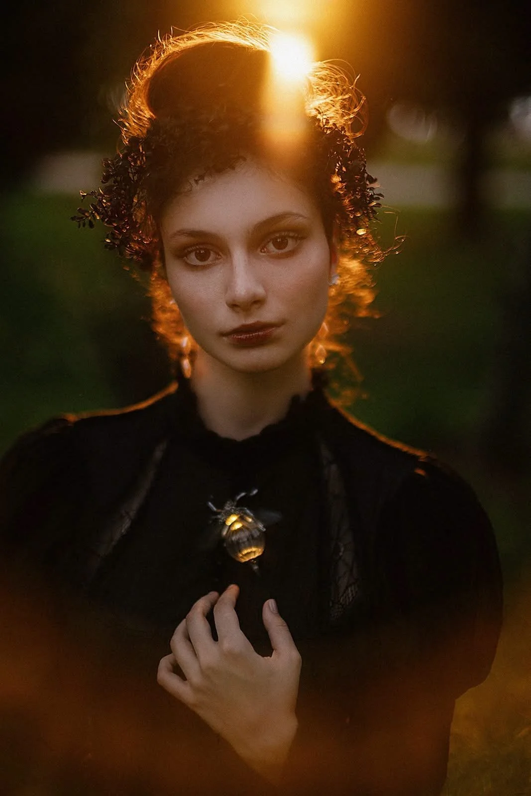 A woman with curly hair and dark makeup standing outside during sunset, holding a bee with a honeycomb near her chest.