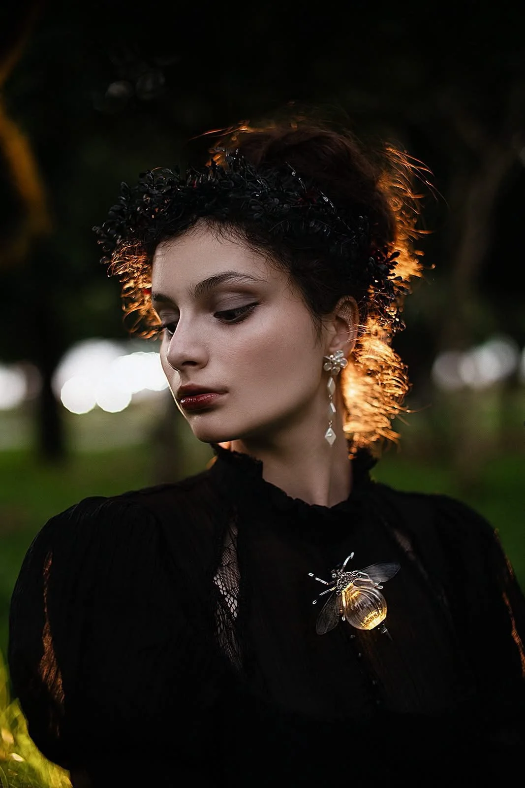 A young woman with dark hair styled with a black floral headband, wearing jewelry including earrings and a brooch shaped like a bee with a gold body, dressed in a black lace outfit, looking down and outdoors at sunset with backlit hair and blurred background.