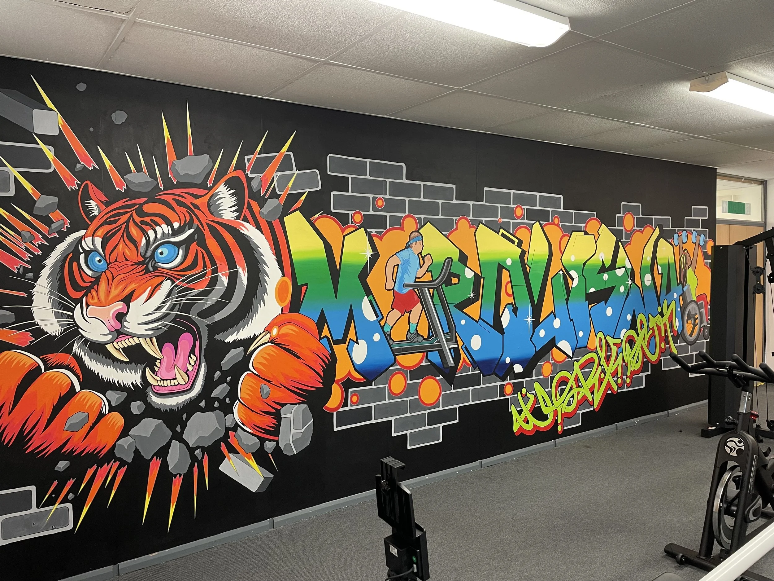 Colorful graffiti mural of a roaring tiger breaking through a wall, with a graffiti-style word on the right side and a runner on a treadmill in front of the mural, all painted on a black wall in a gym.