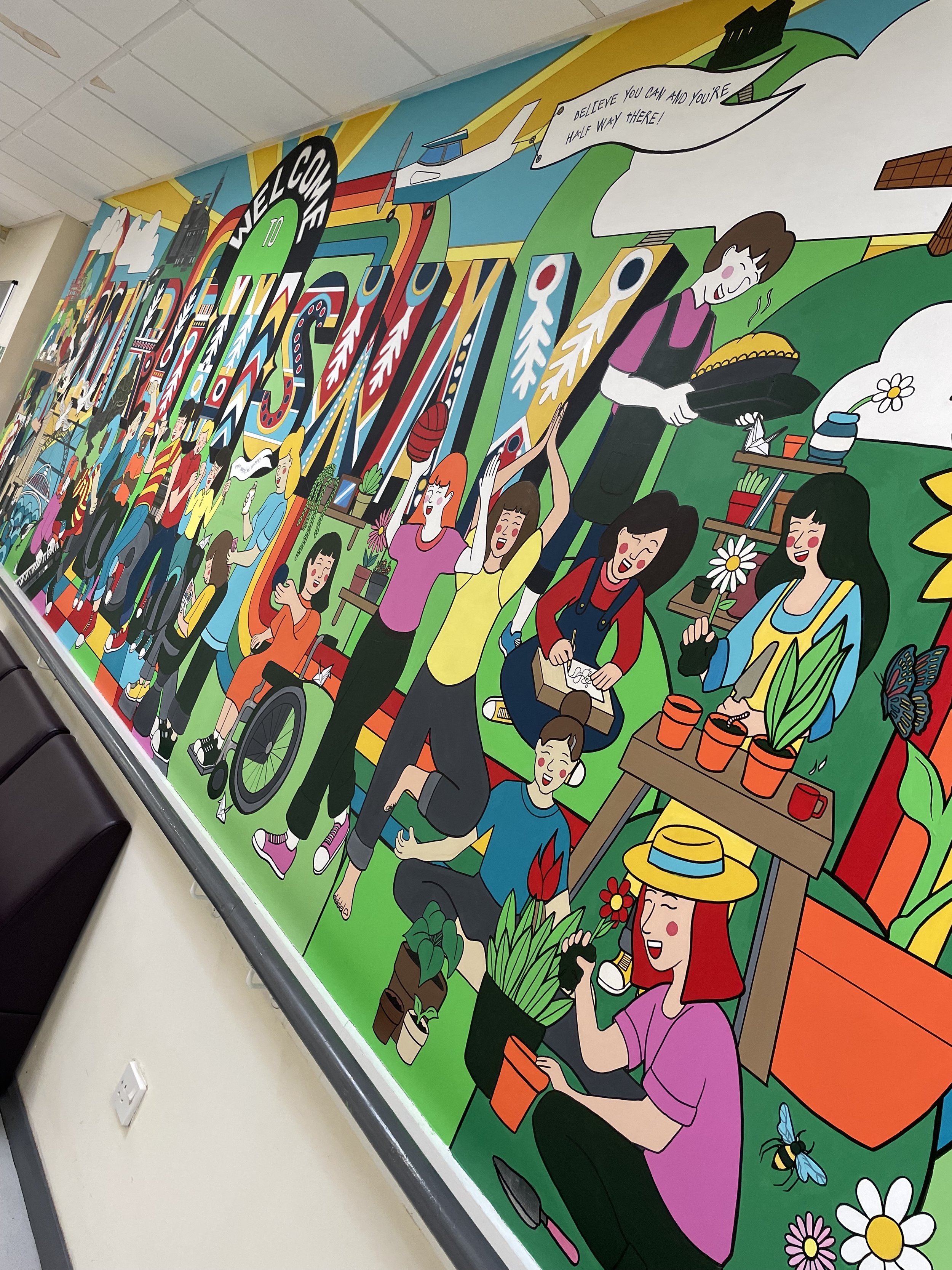 Colorful mural depicting diverse people engaged in activities such as planting, gardening, cooking, painting, and socializing, with scenic background elements like hills, flowers, and clouds.