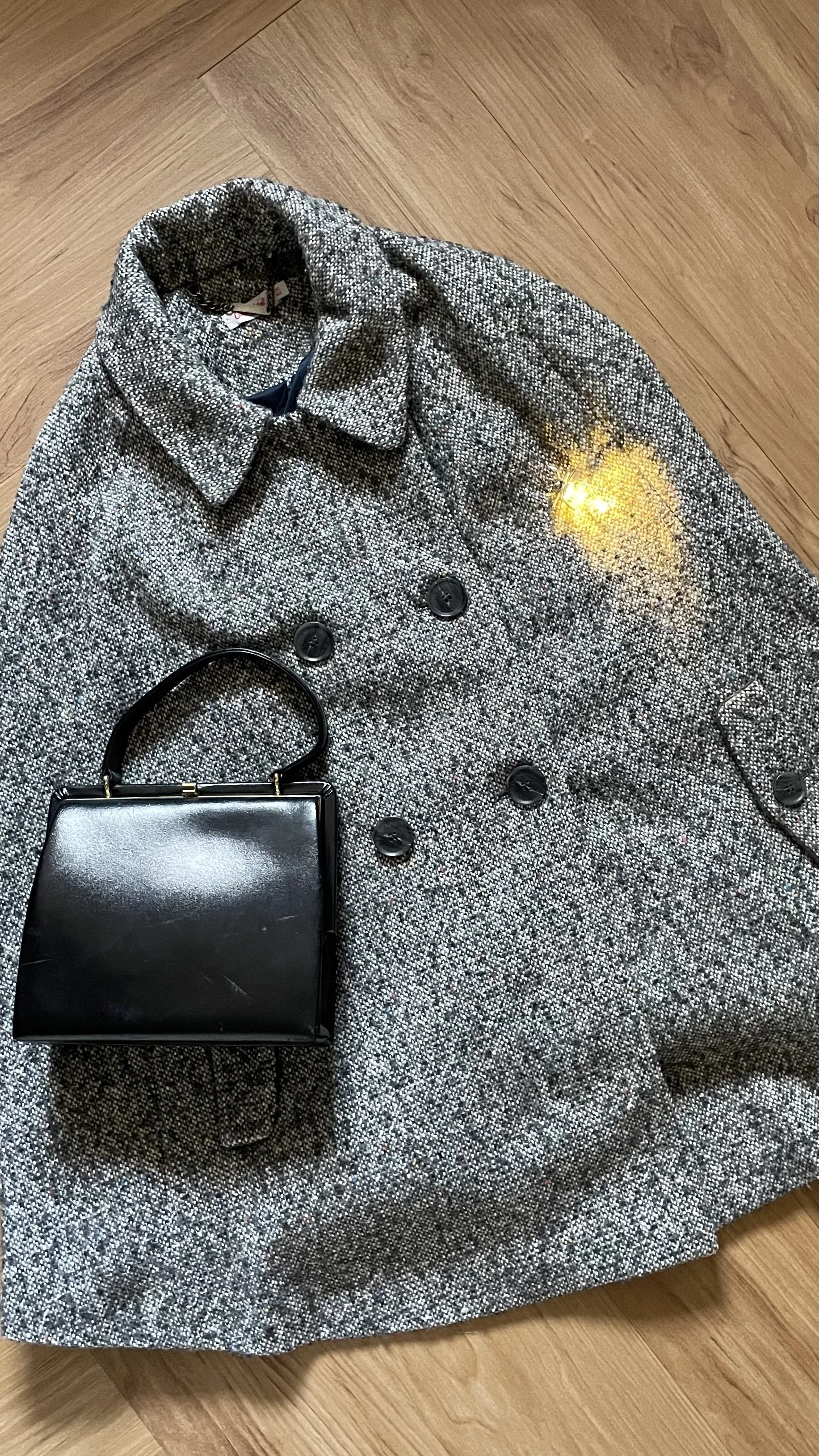 A gray textured coat, a small black handbag, and a bright yellow light are on a wooden floor.
