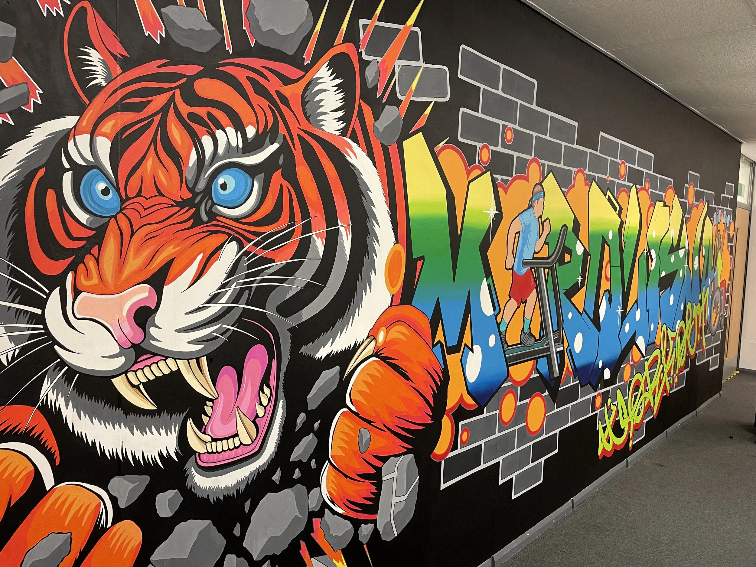Colorful graffiti mural featuring a fierce tiger emerging from the wall, with bright blue eyes, sharp teeth, and orange, black, and white fur. The mural also depicts a runner on a treadmill and large, vibrant, overlapping graffiti letters, with a bac
