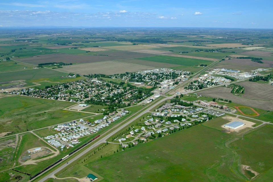 An aerial view of a small town surrounded by farmland, with residential areas, a highway running through, and open green fields.