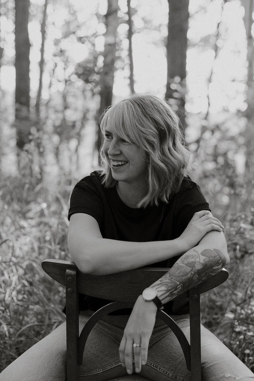 A smiling woman with shoulder-length wavy hair, wearing a black shirt, sitting outdoors in a forested area, resting her arms on a chair with a tattooed arm visible, in black and white
