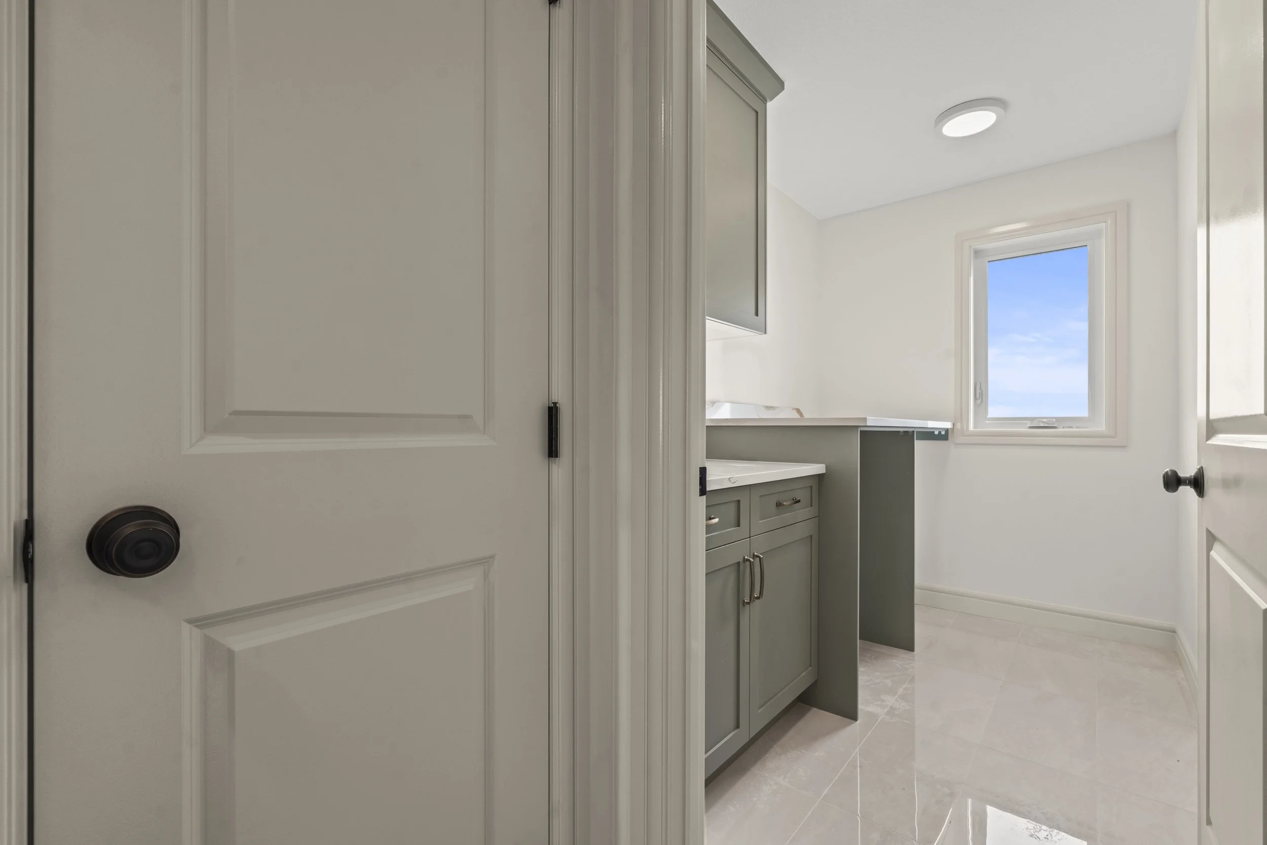 A laundry room with beige tiled floor, white walls, a window showing a blue sky, grey cabinets, and a white countertop.