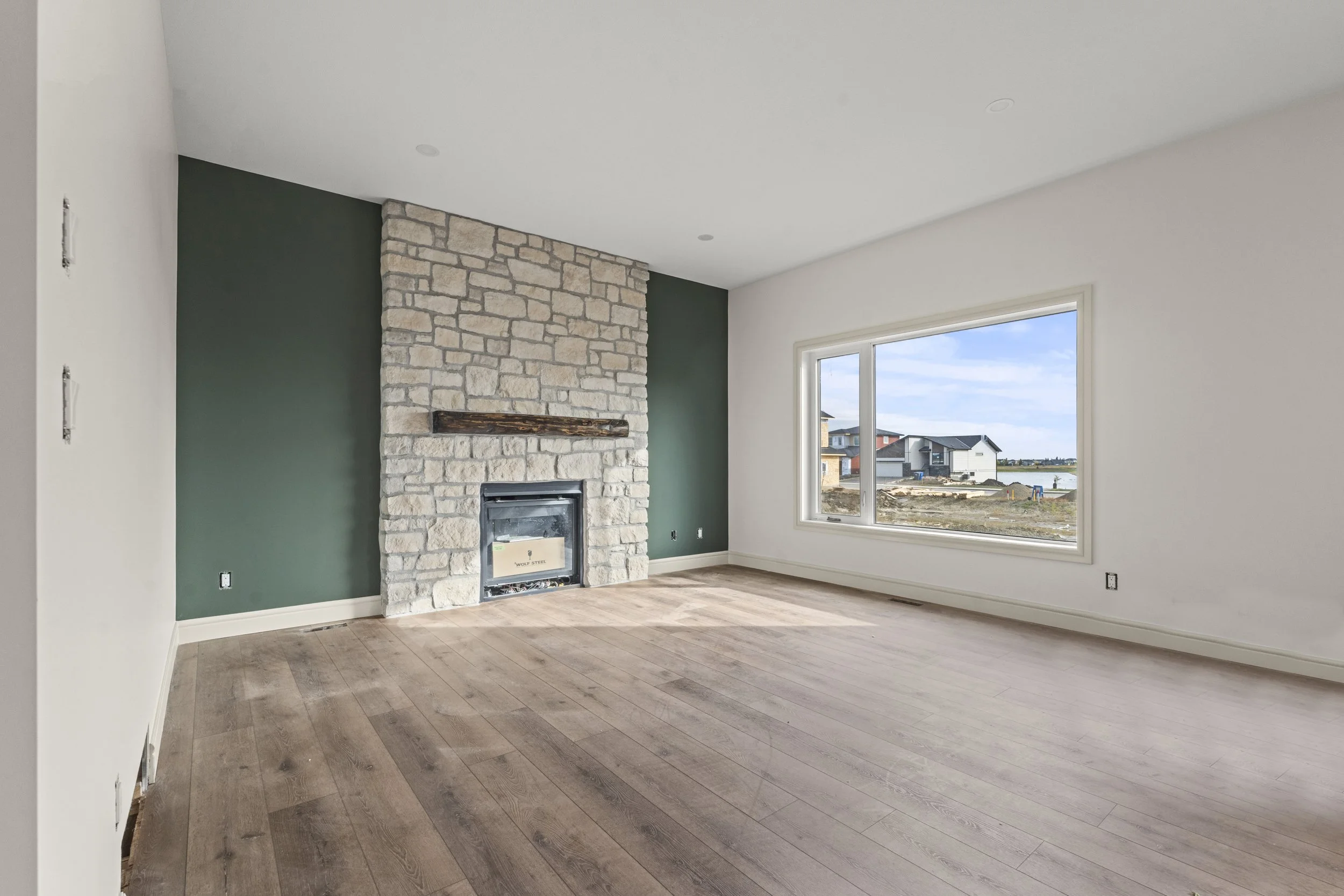 Empty living room with wooden floor, stone fireplace, green accent wall, and large window showing houses outside.