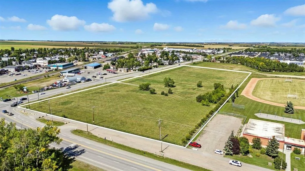 An aerial view of an empty grassy lot surrounded by roads and nearby buildings, with a sports field and park on the right, and commercial area in the background under a partly cloudy sky.