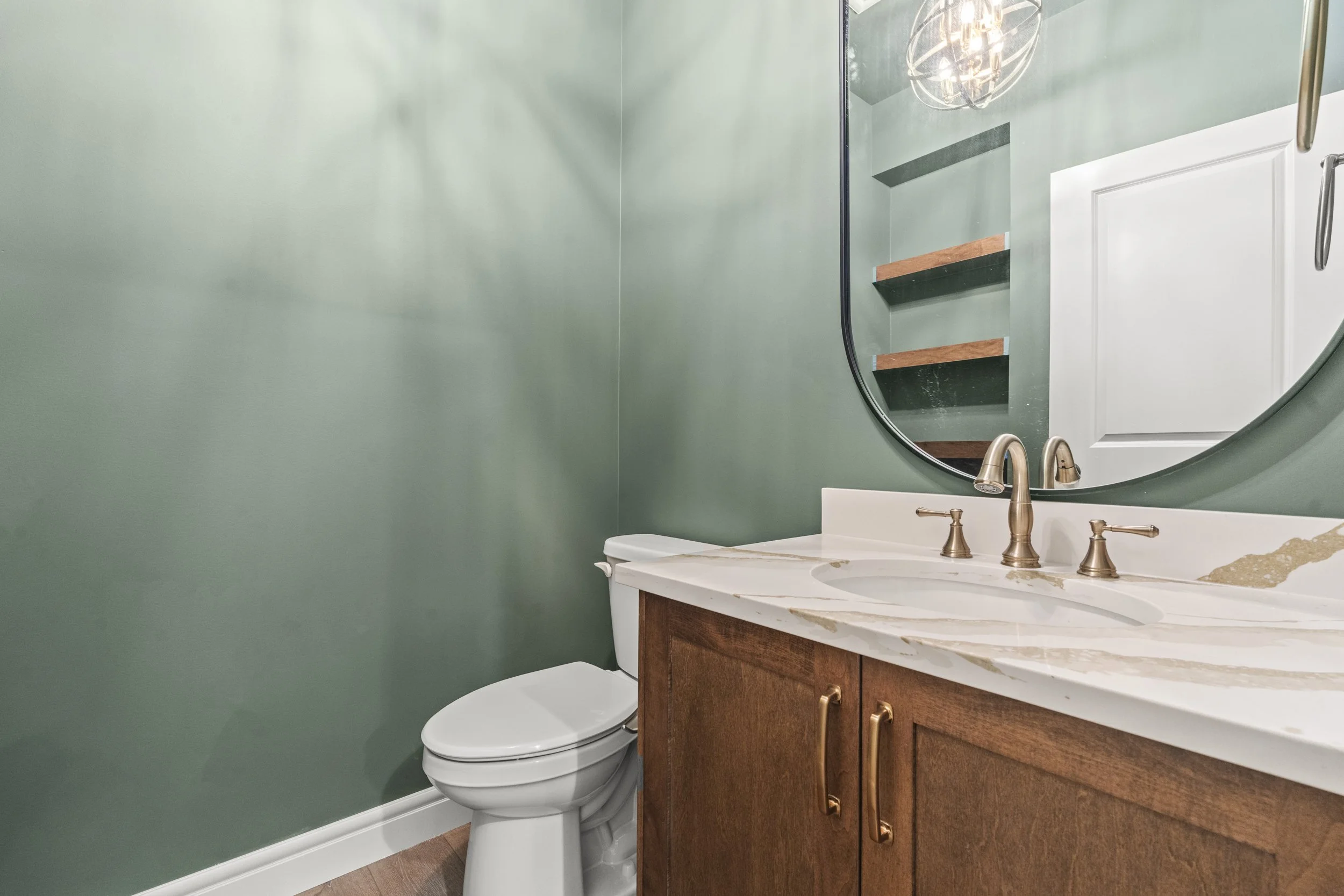 Bathroom with olive green wall, white toilet, wooden vanity cabinet, marble countertop, oval mirror, brushed gold faucet, and open shelves reflected in mirror.