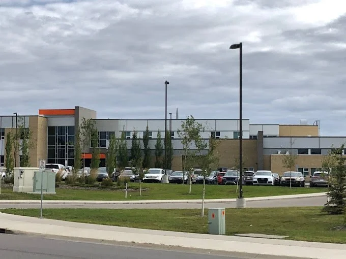 Modern hospital building with parking lot full of vehicles, surrounded by trees and street lamps, under a cloudy sky.