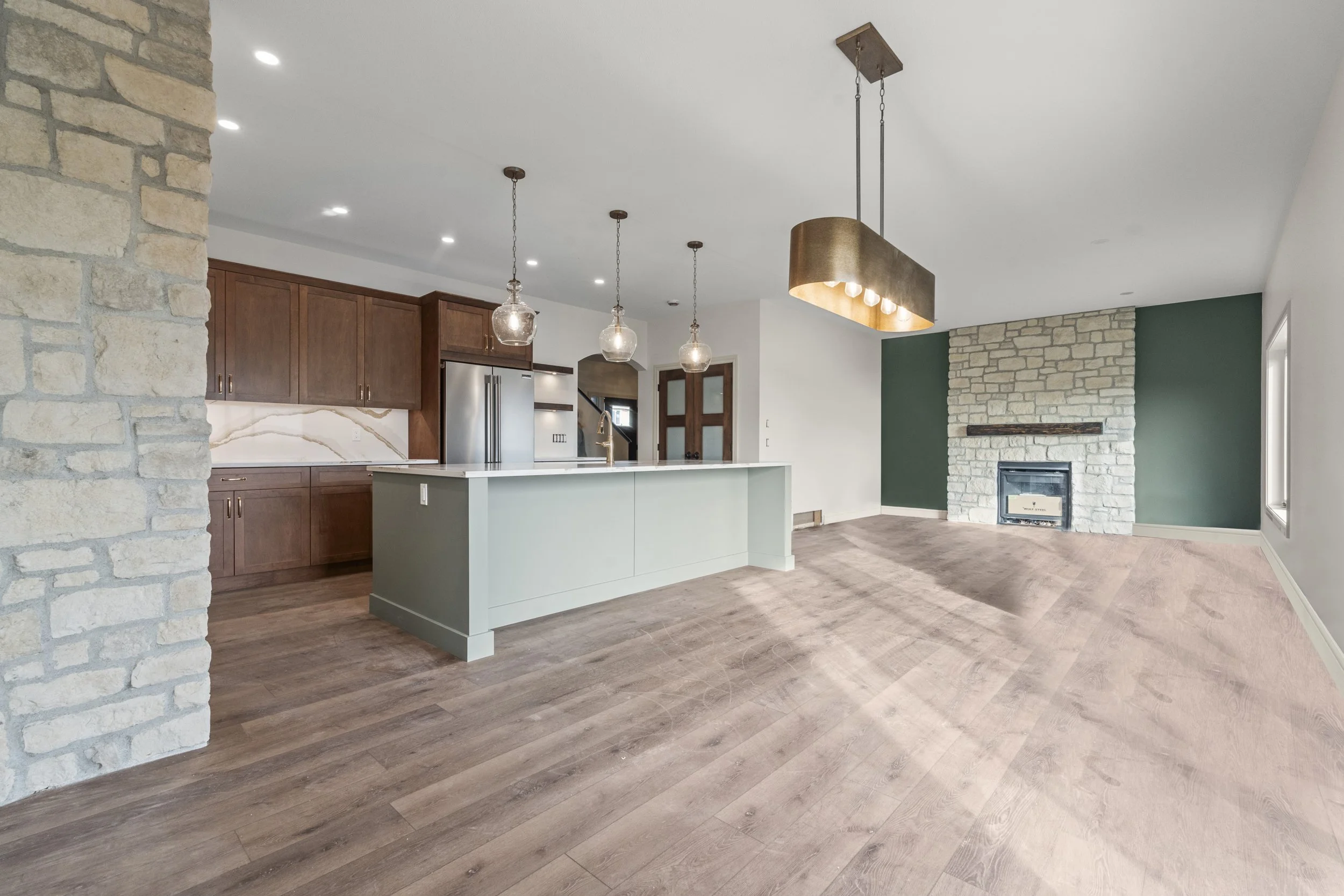 Open-concept living space with a kitchen featuring wooden cabinets, white marble backsplash, and a light-colored island. The room has hardwood floors, a green accent wall with a white stone fireplace, and modern pendant lights hanging over the island