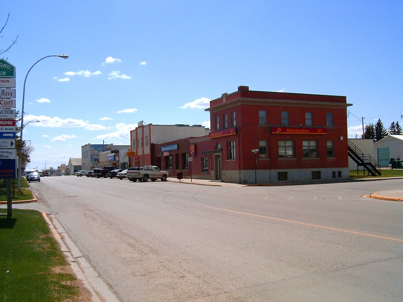 A small town street scene under a bright blue sky with some clouds. There is a red brick building with an LED sign reading 'CIBC Banking Centre' on the corner. Several other storefronts and parked cars are along the street.