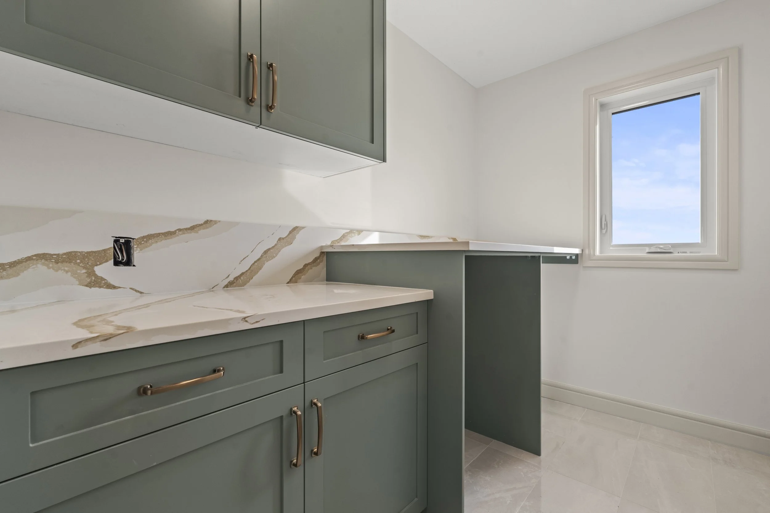 Empty kitchen with green cabinets, marble countertops, a marble backsplash, a window showing blue sky, and an electrical outlet without a cover.